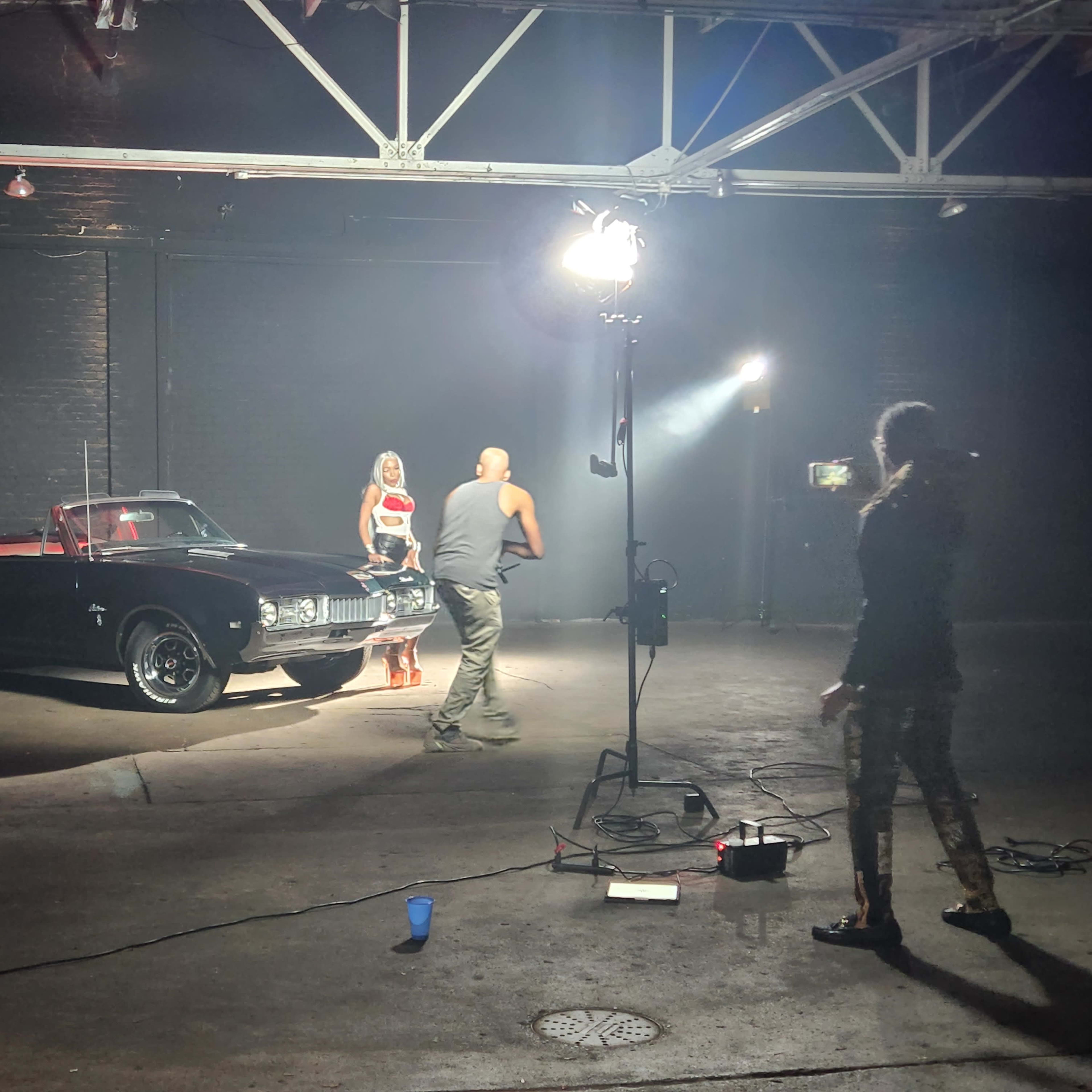 A woman in a red and white outfit stands near a black convertible car while a man interacts with her in a dimly lit warehouse.