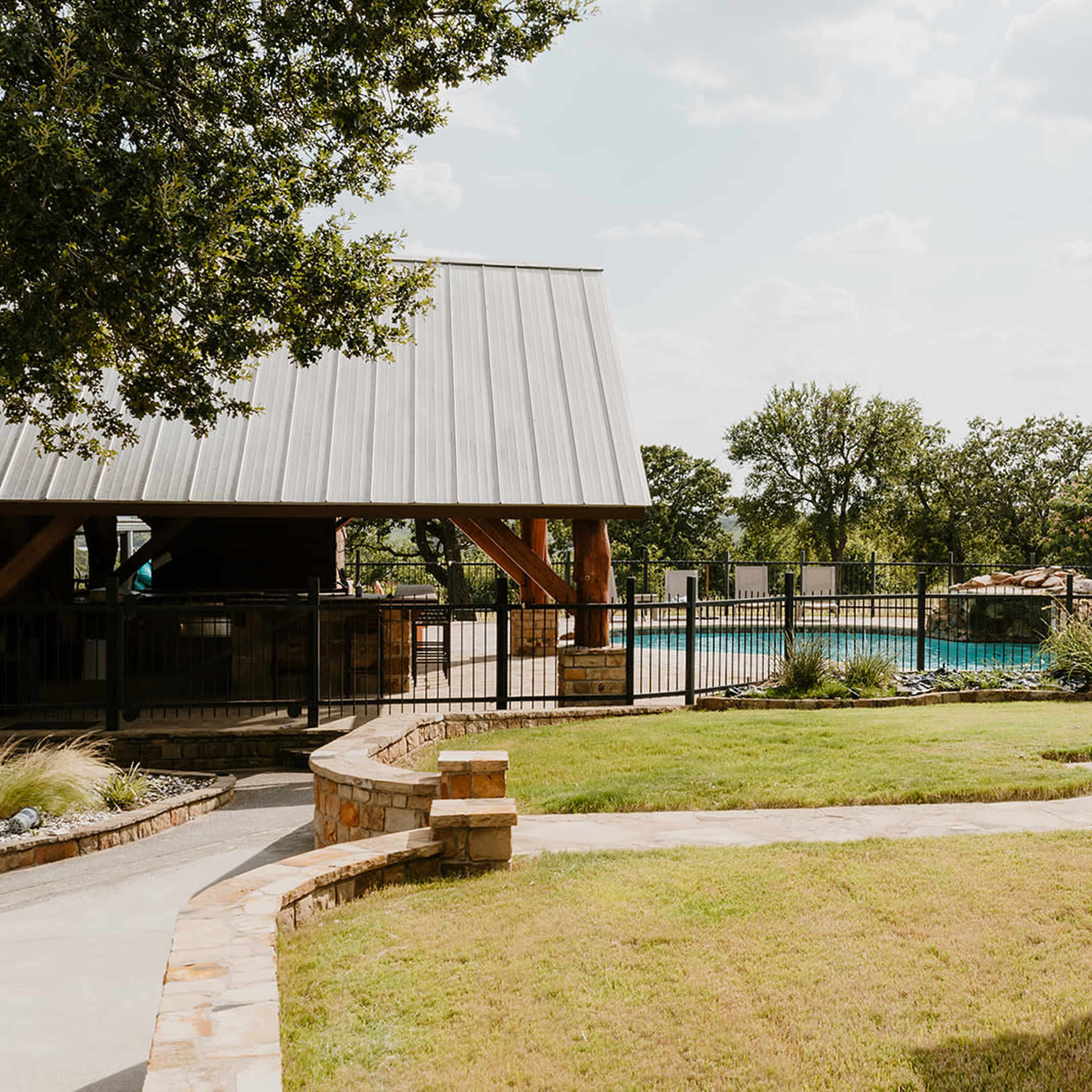 A spacious outdoor area with a fenced swimming pool, surrounded by greenery and a nearby covered structure.