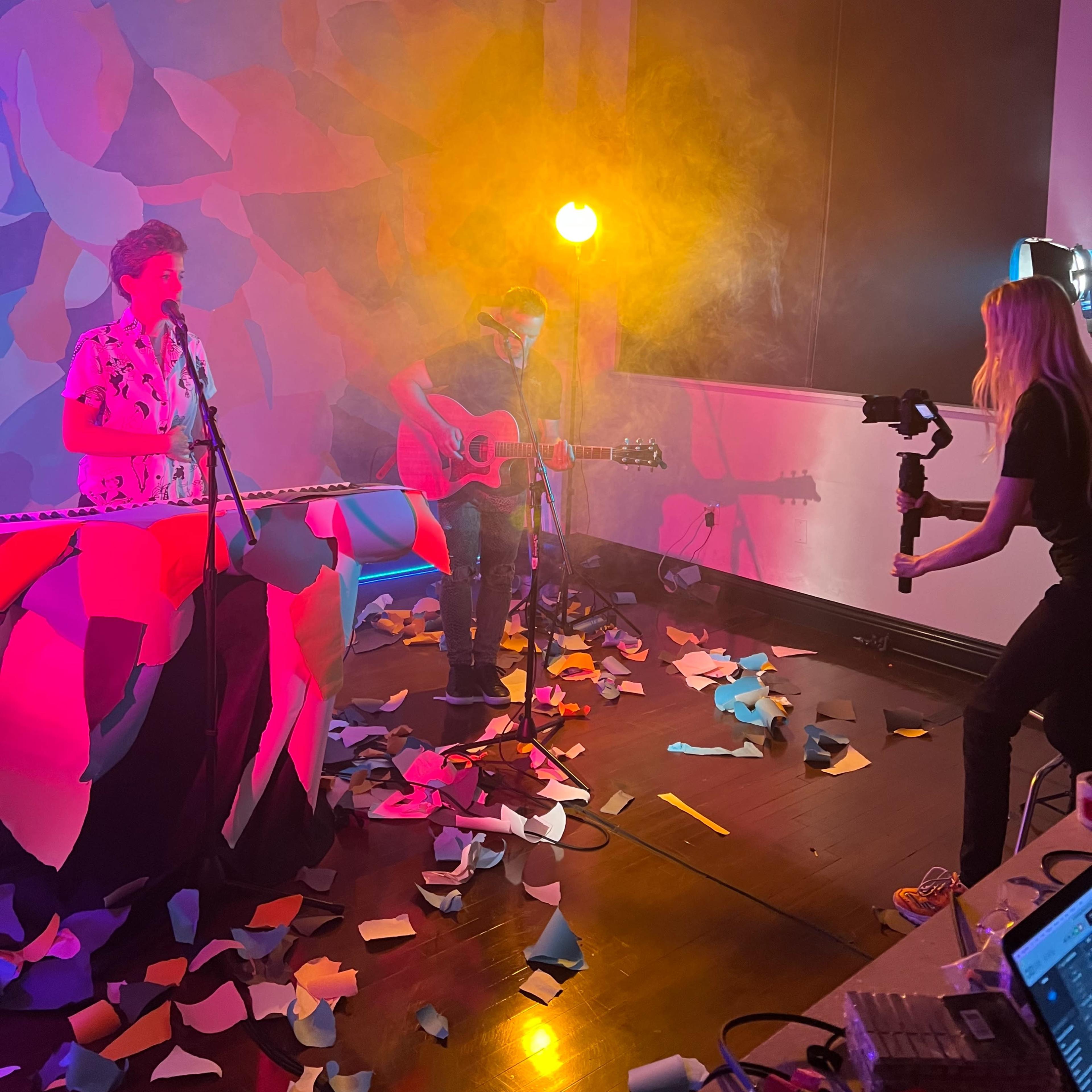 A musician plays guitar while a singer performs, surrounded by colorful paper scattered on the floor and an illuminated backdrop.