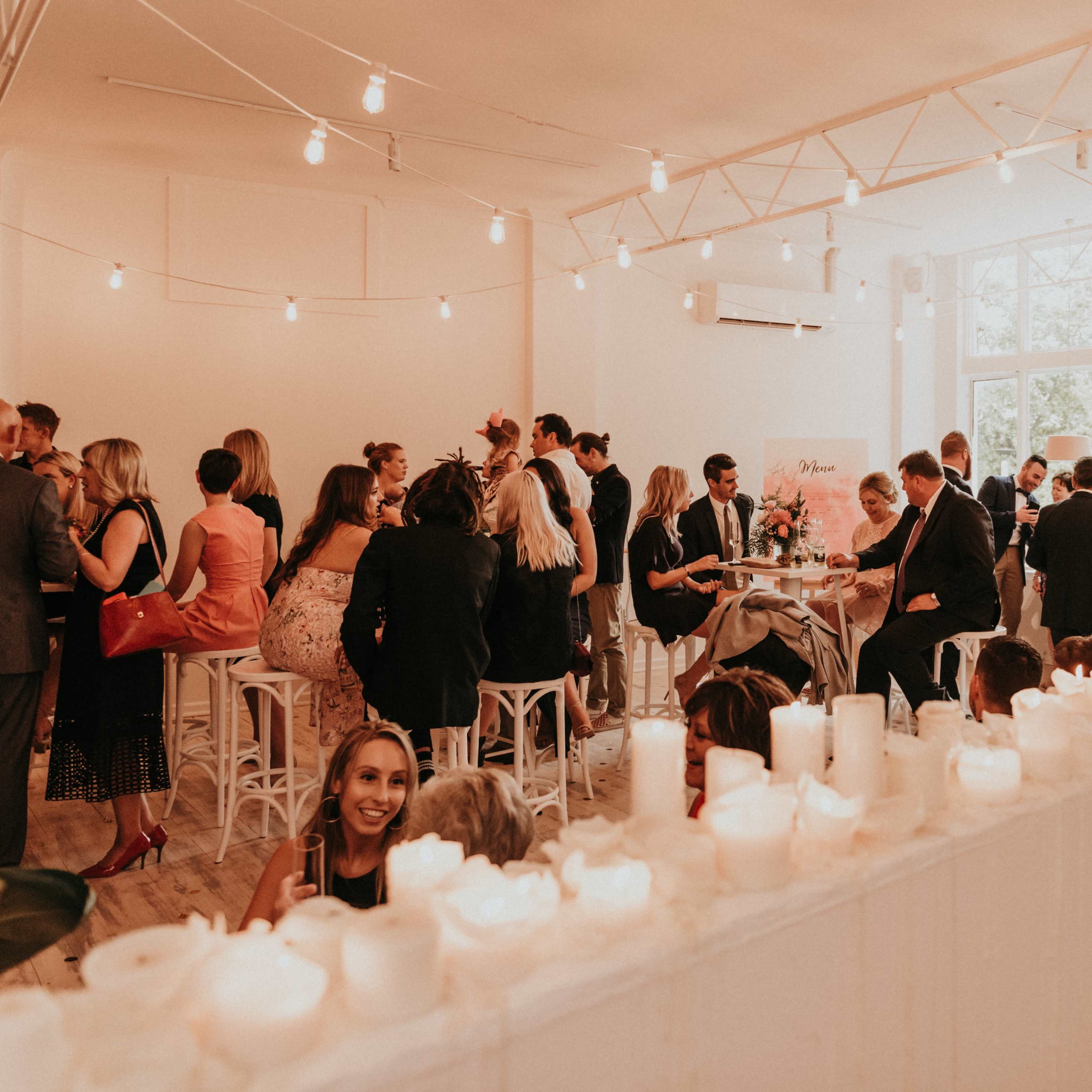 A lively gathering takes place in a brightly lit, modern venue, with guests mingling and seated around tables adorned with candles.