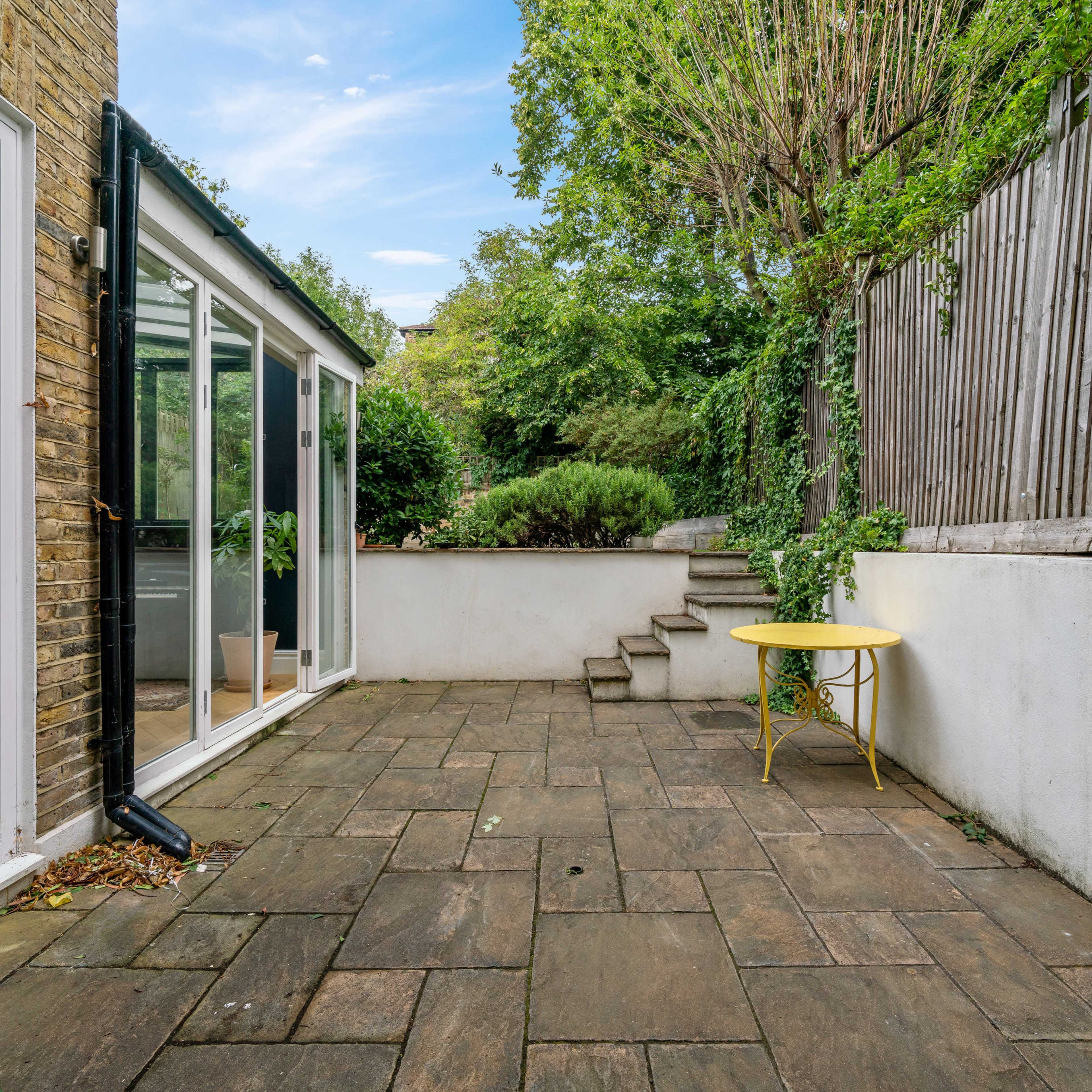 A stone-paved outdoor area with a yellow table, bordered by a glass structure and a wooden fence, surrounded by greenery.