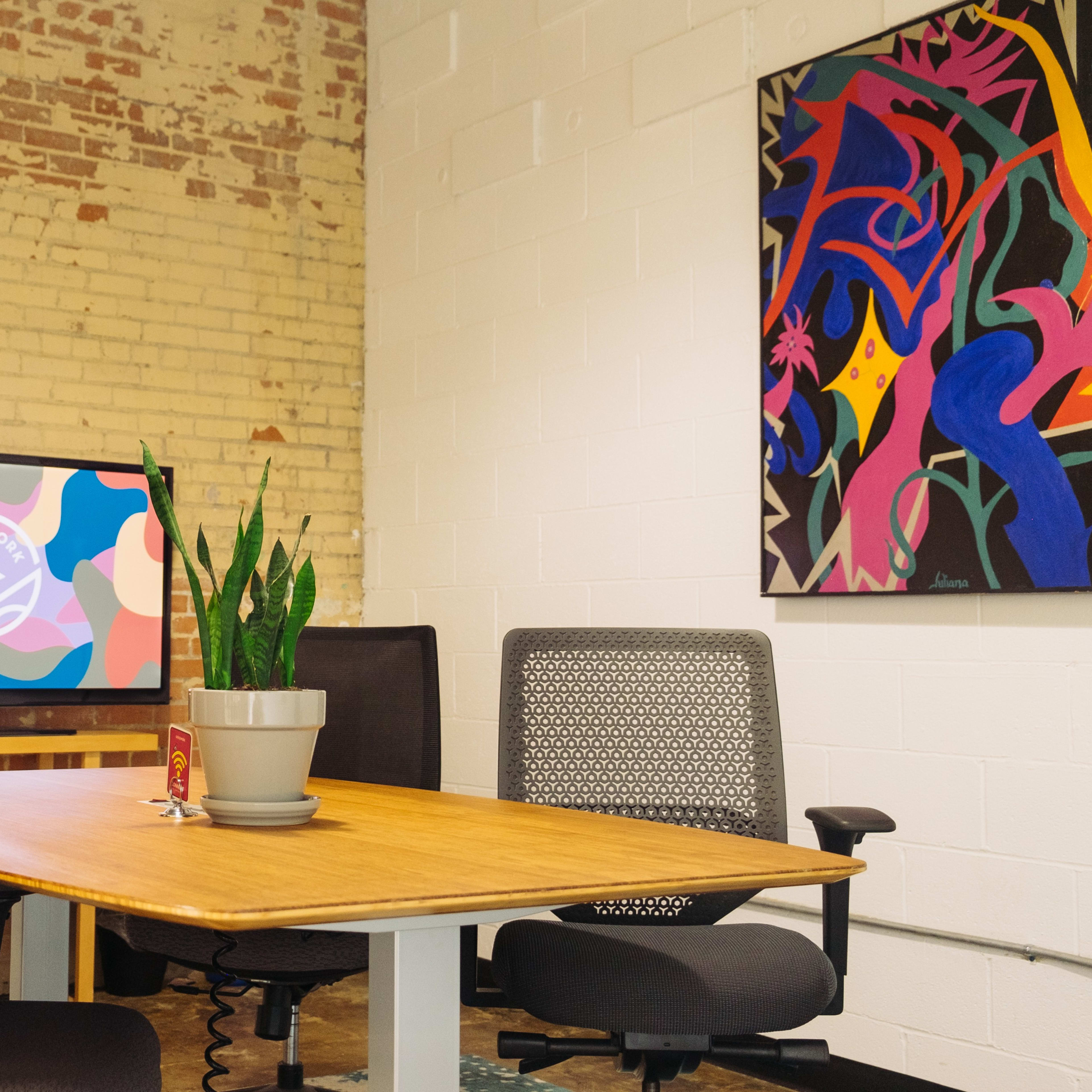 A conference room features a wooden table, four chairs, a potted plant, and a wall-mounted screen displaying a colorful graphic, with a vibrant artwork on an adjacent wall.