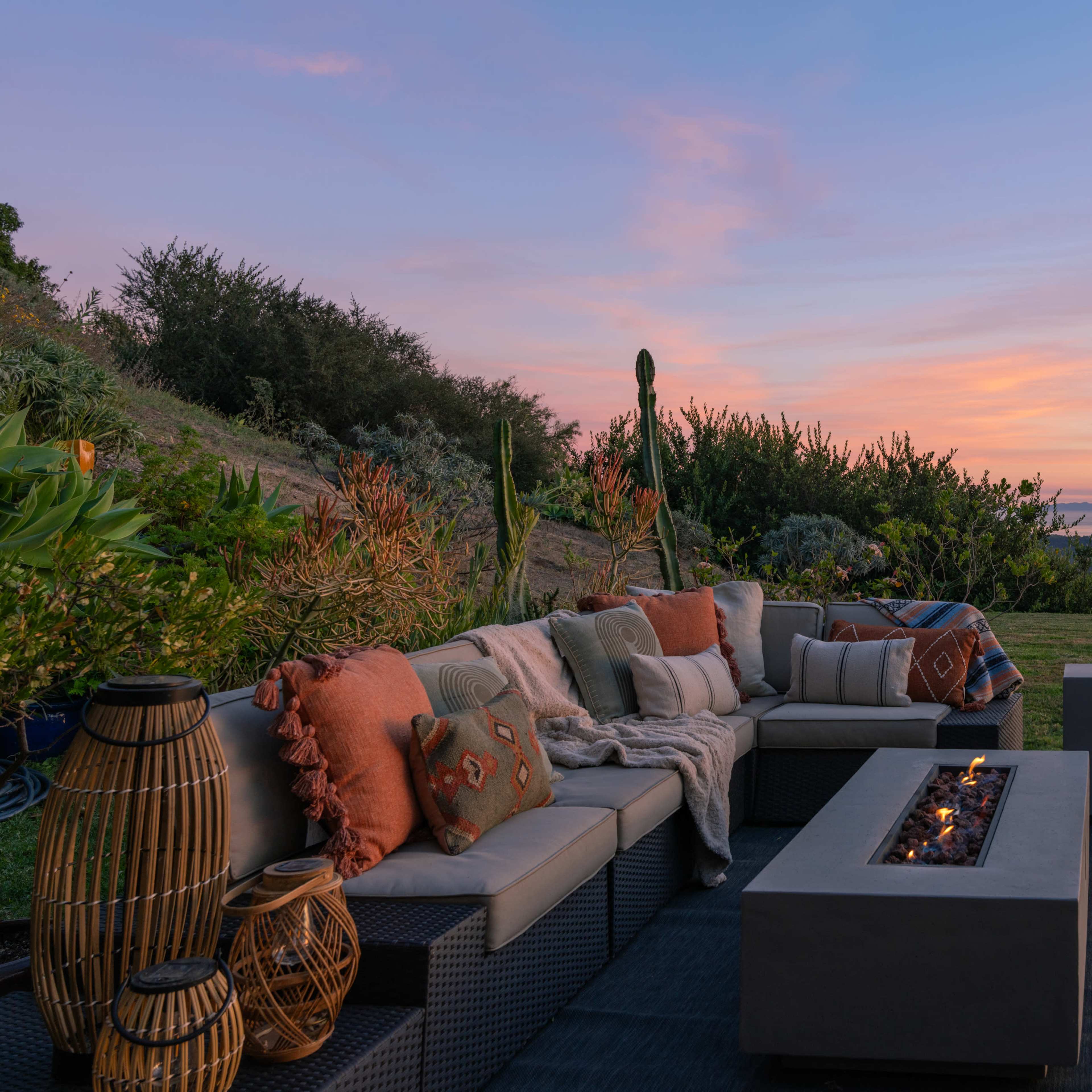 A spacious outdoor lounge area features a modern fire pit surrounded by comfortable seating, with a view of the sunset over the landscape.
