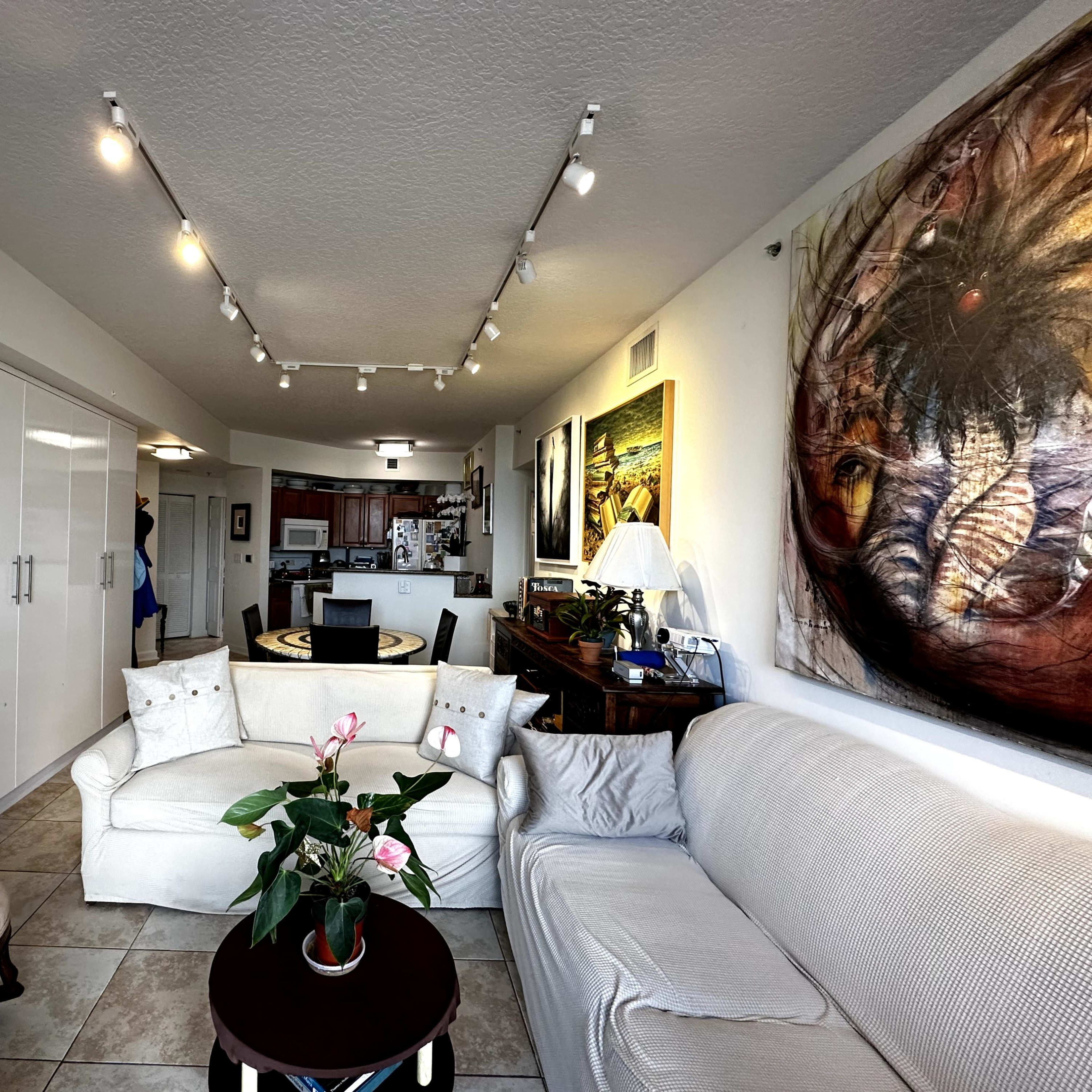 A modern living room with a white sofa, an armchair, and a dining area visible in the background, along with wall art and cabinetry.
