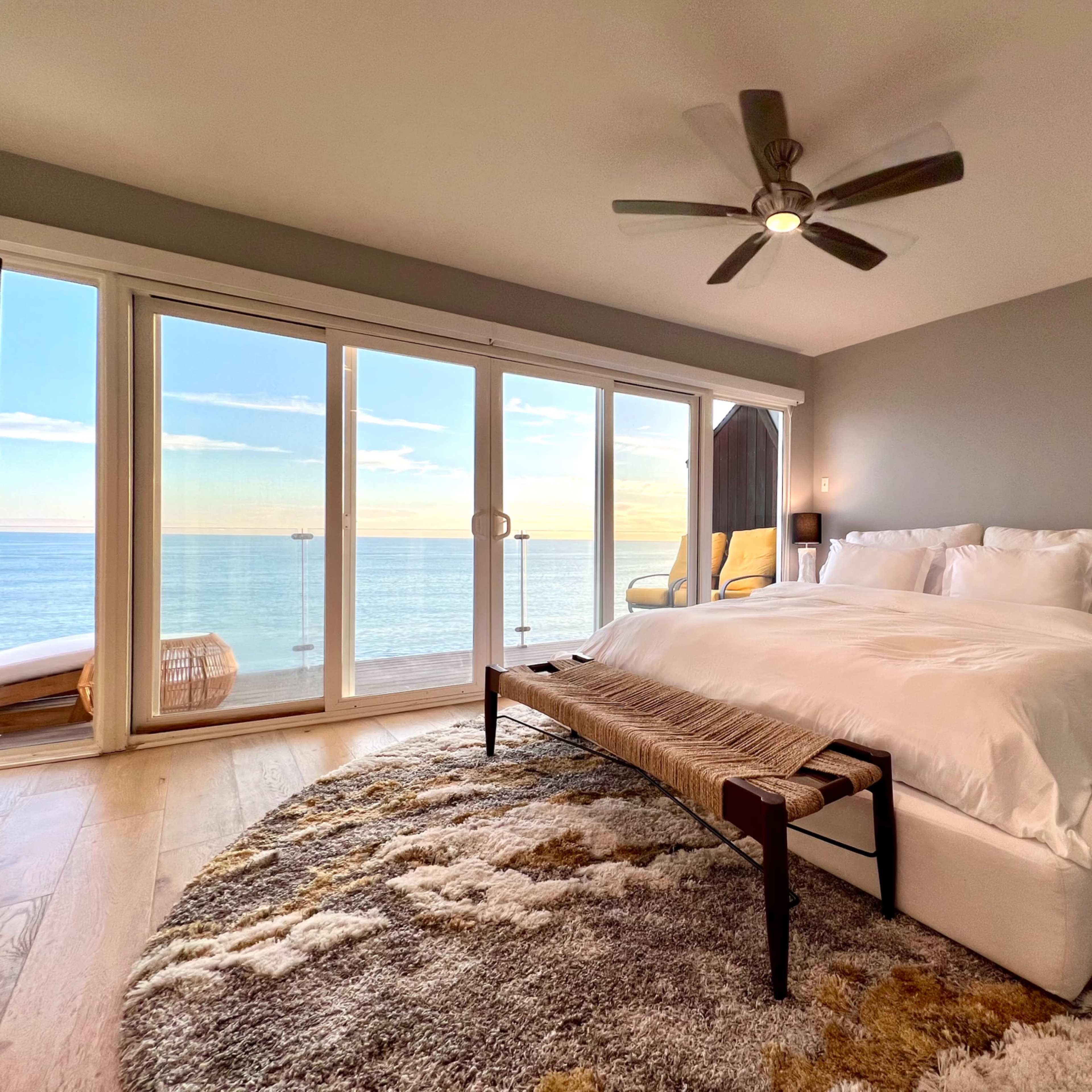 A modern bedroom features a large bed, a patterned area rug, and floor-to-ceiling windows overlooking the ocean.