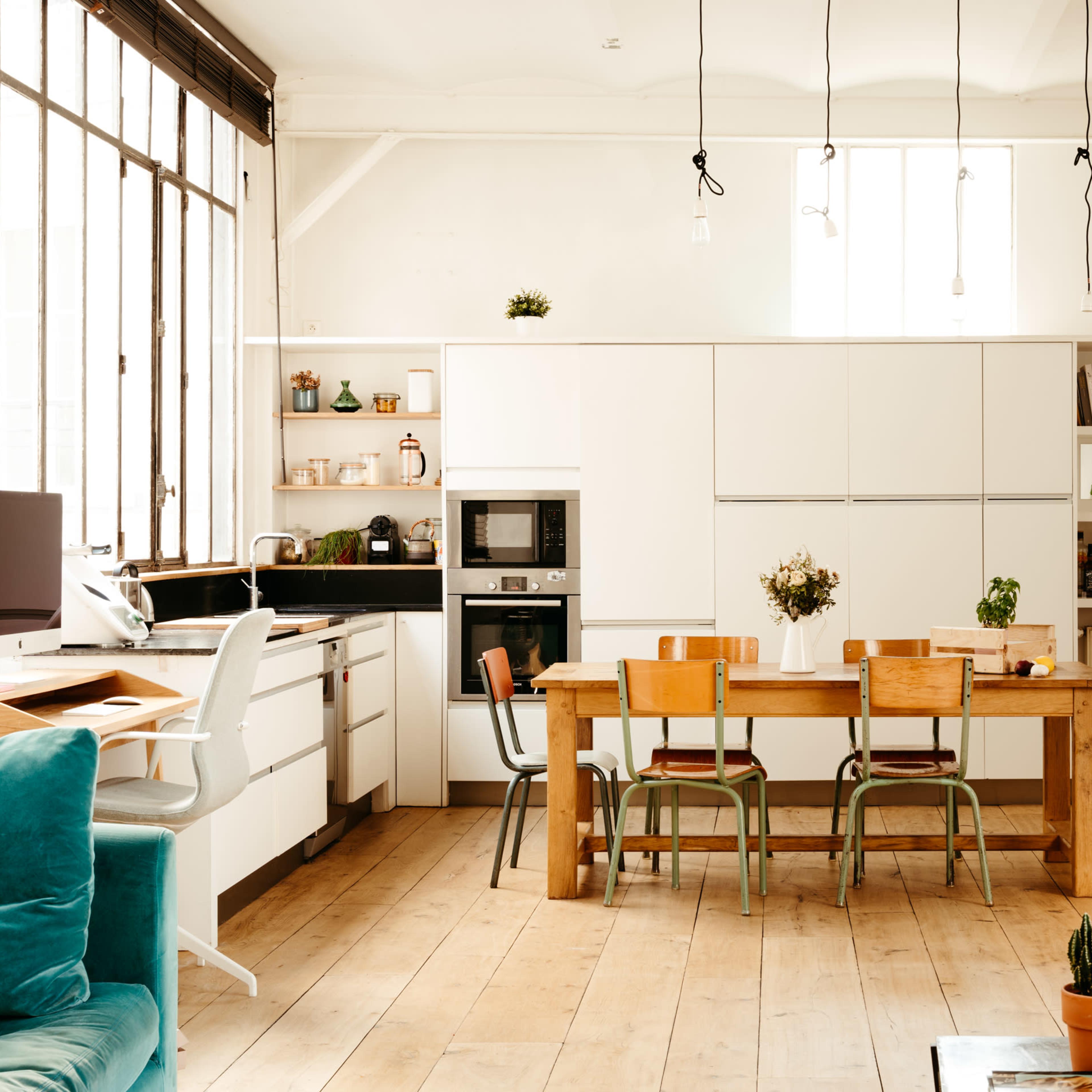 A modern kitchen and dining area features a wooden table surrounded by chairs, with white cabinetry, appliances, and large windows allowing natural light to fill the space.