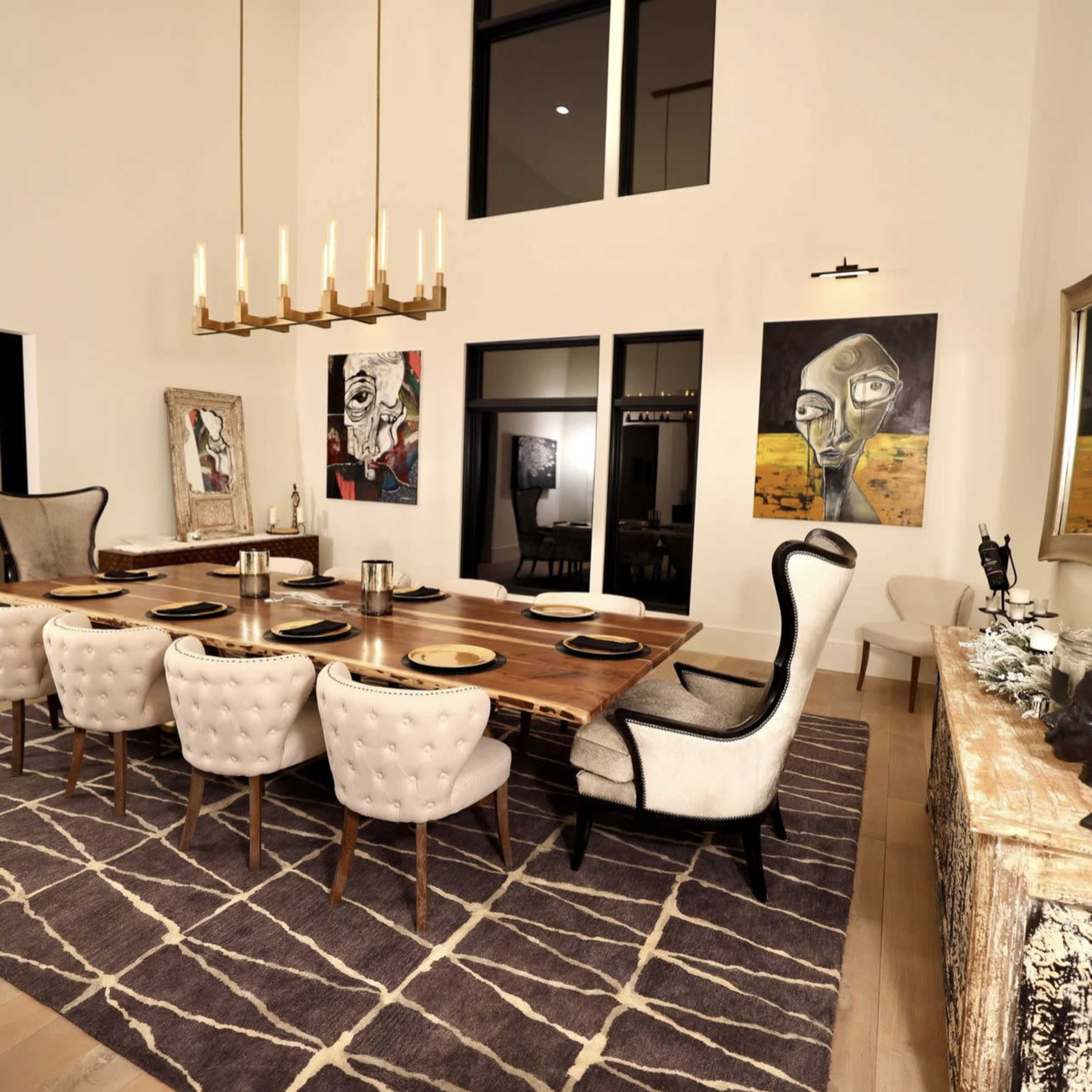 A modern dining room features a long wooden table with upholstered chairs, surrounded by artwork on the walls and illuminated by a chandelier.
