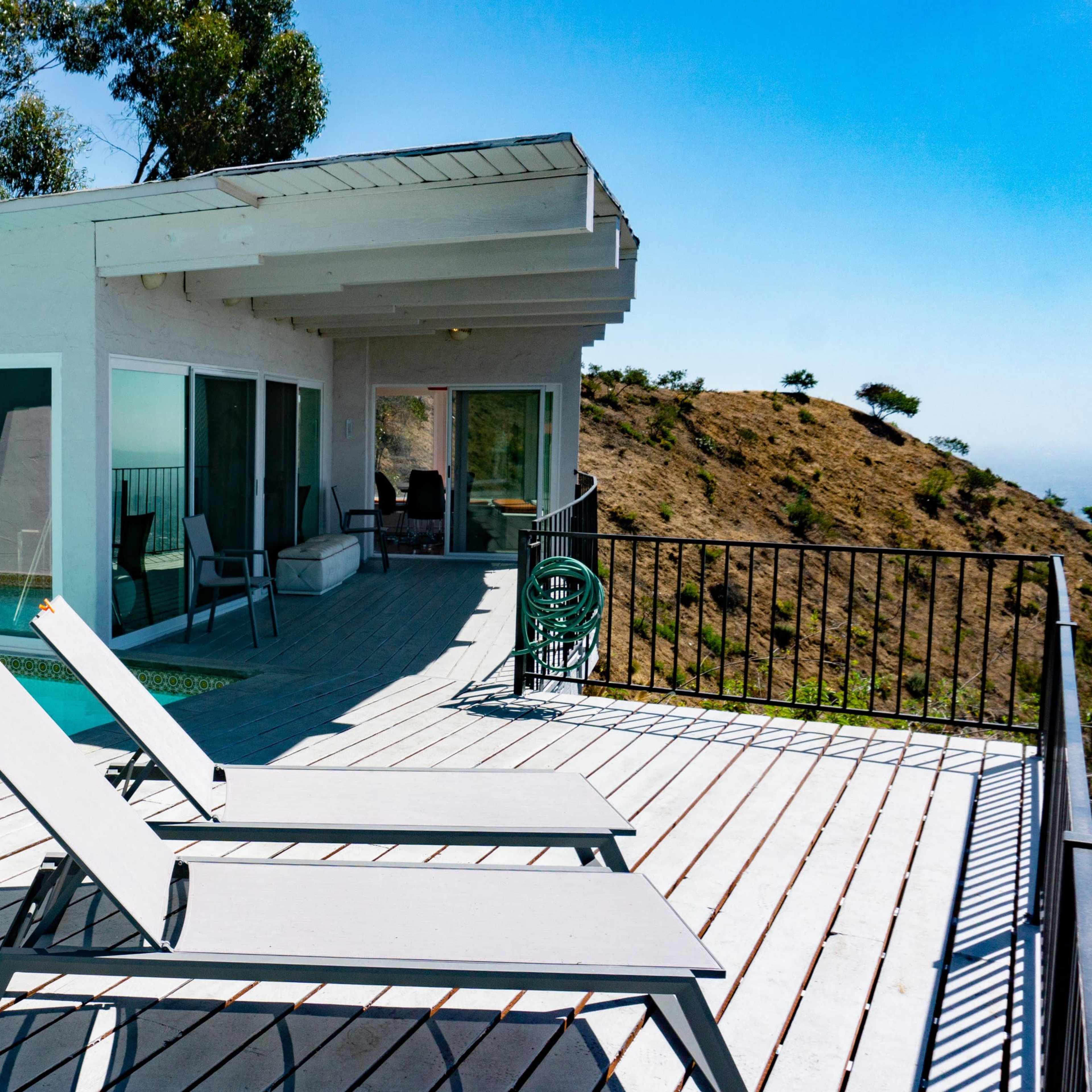 A modern hillside home features a balcony with poolside lounge chairs overlooking a natural landscape.