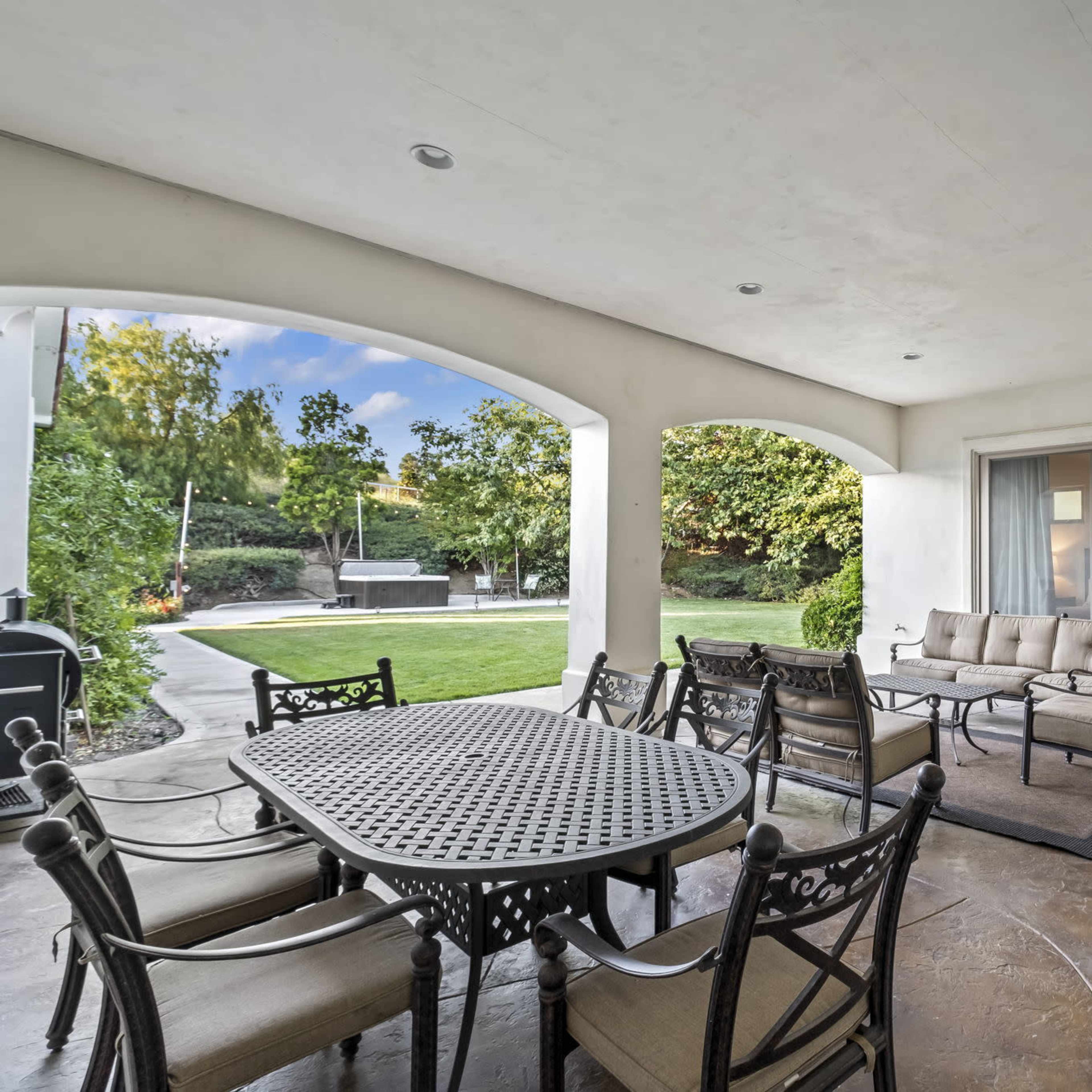 A spacious outdoor patio with a dining table and seating area adjacent to a well-manicured lawn and a pool.