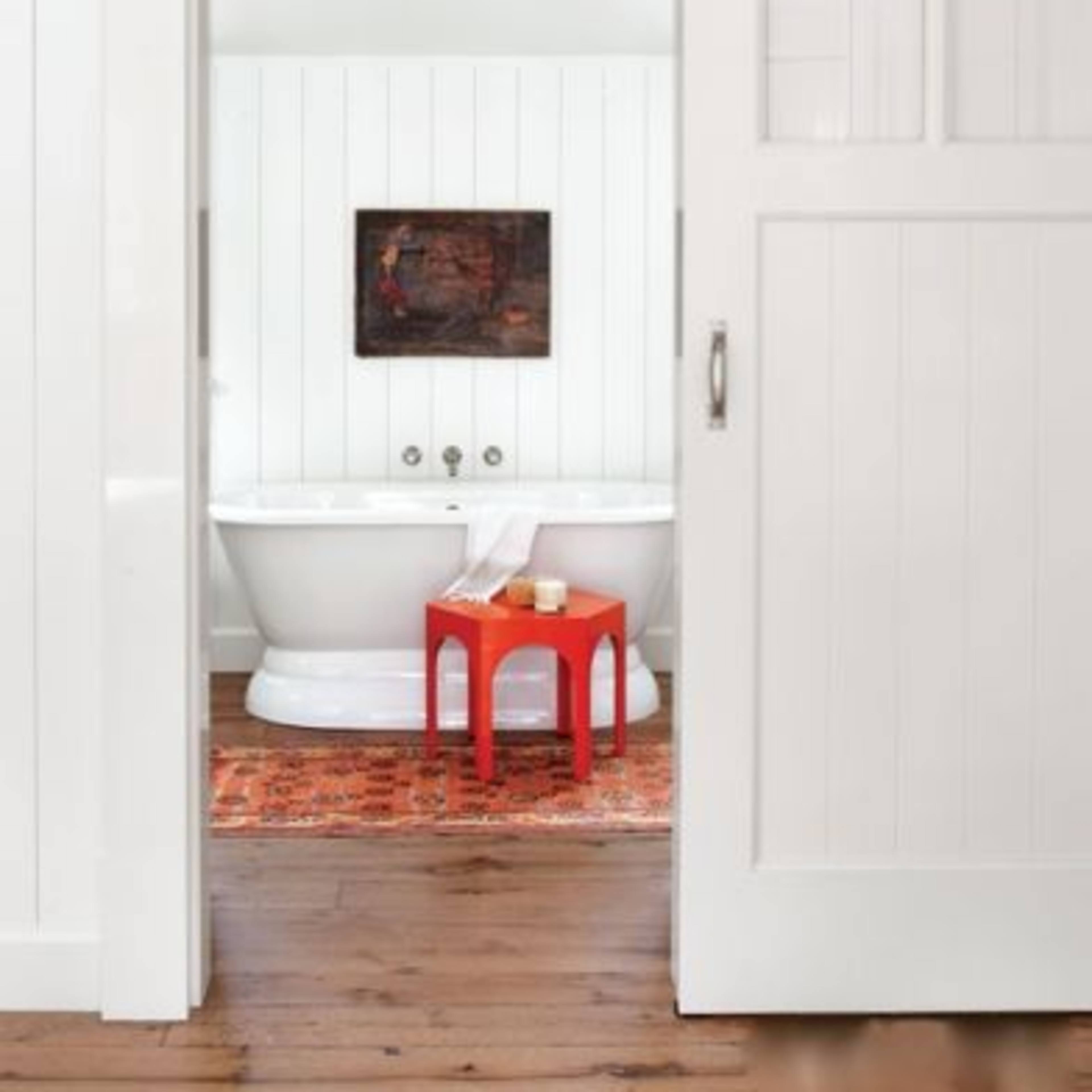 A white-walled bathroom with a freestanding bathtub, a small red table, and a rug on a wooden floor.