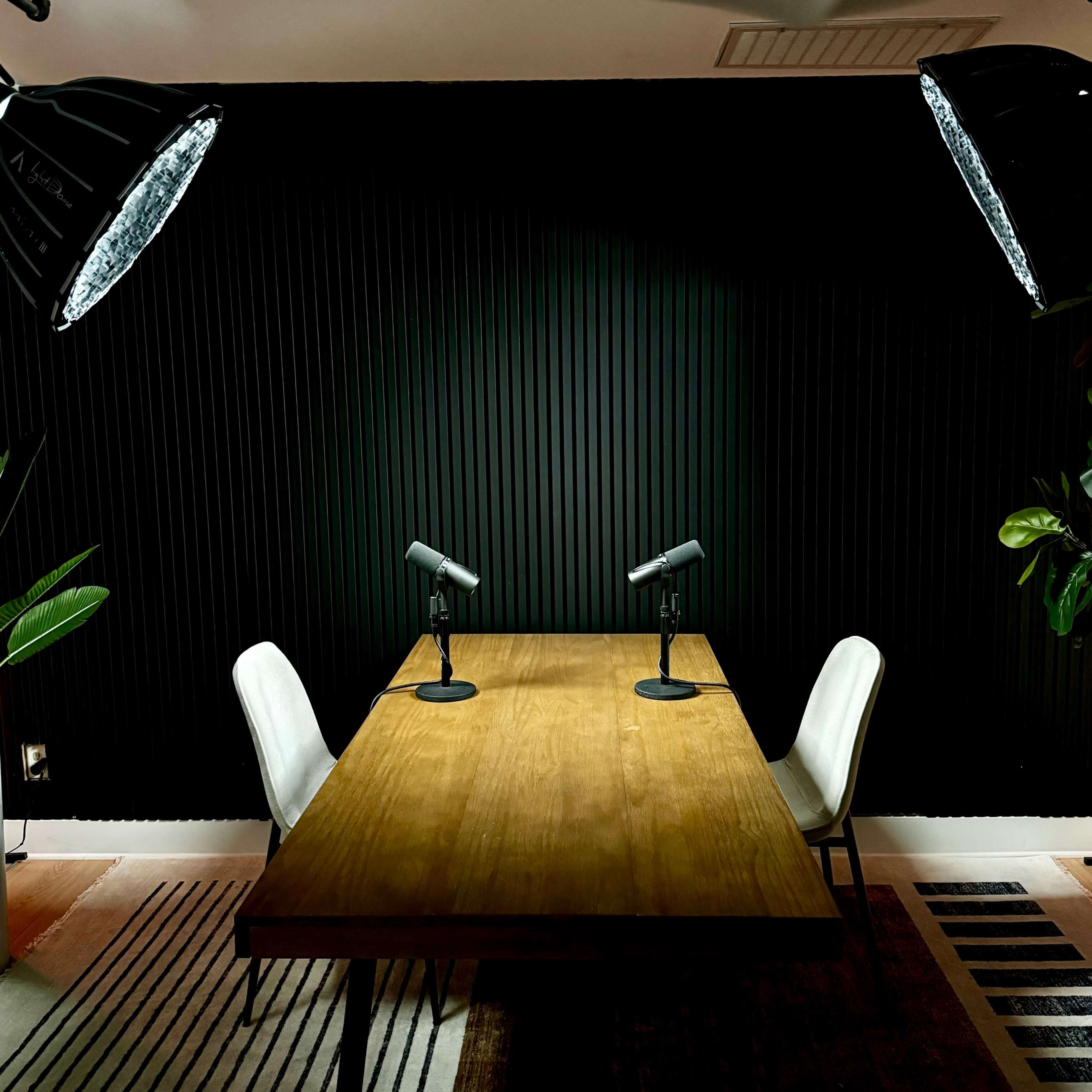 A wooden table with two chairs and microphones is set under bright lights against a dark, ribbed wall.