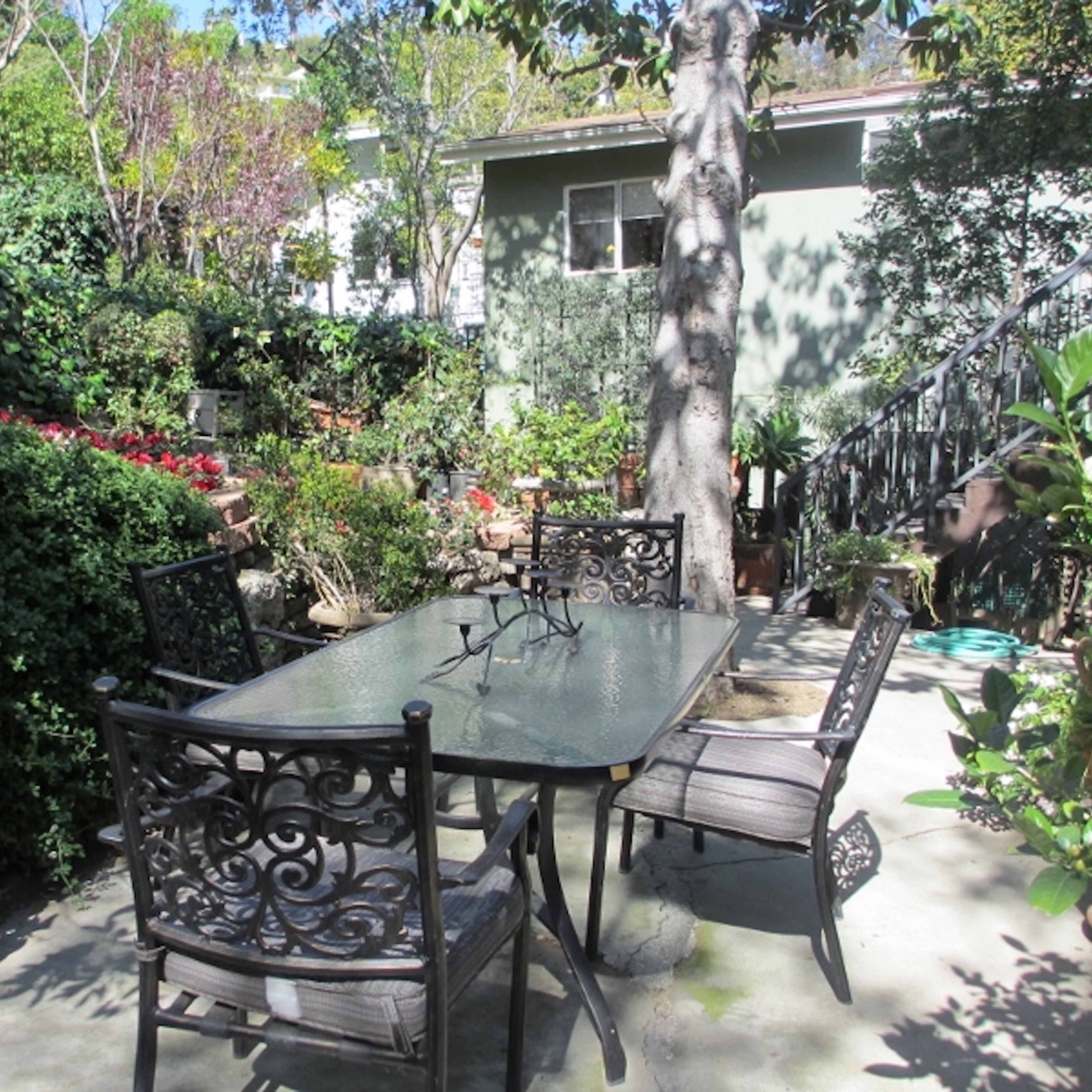 A set of four wrought iron chairs surrounds a glass-top table in a lush garden filled with greenery and flowers.