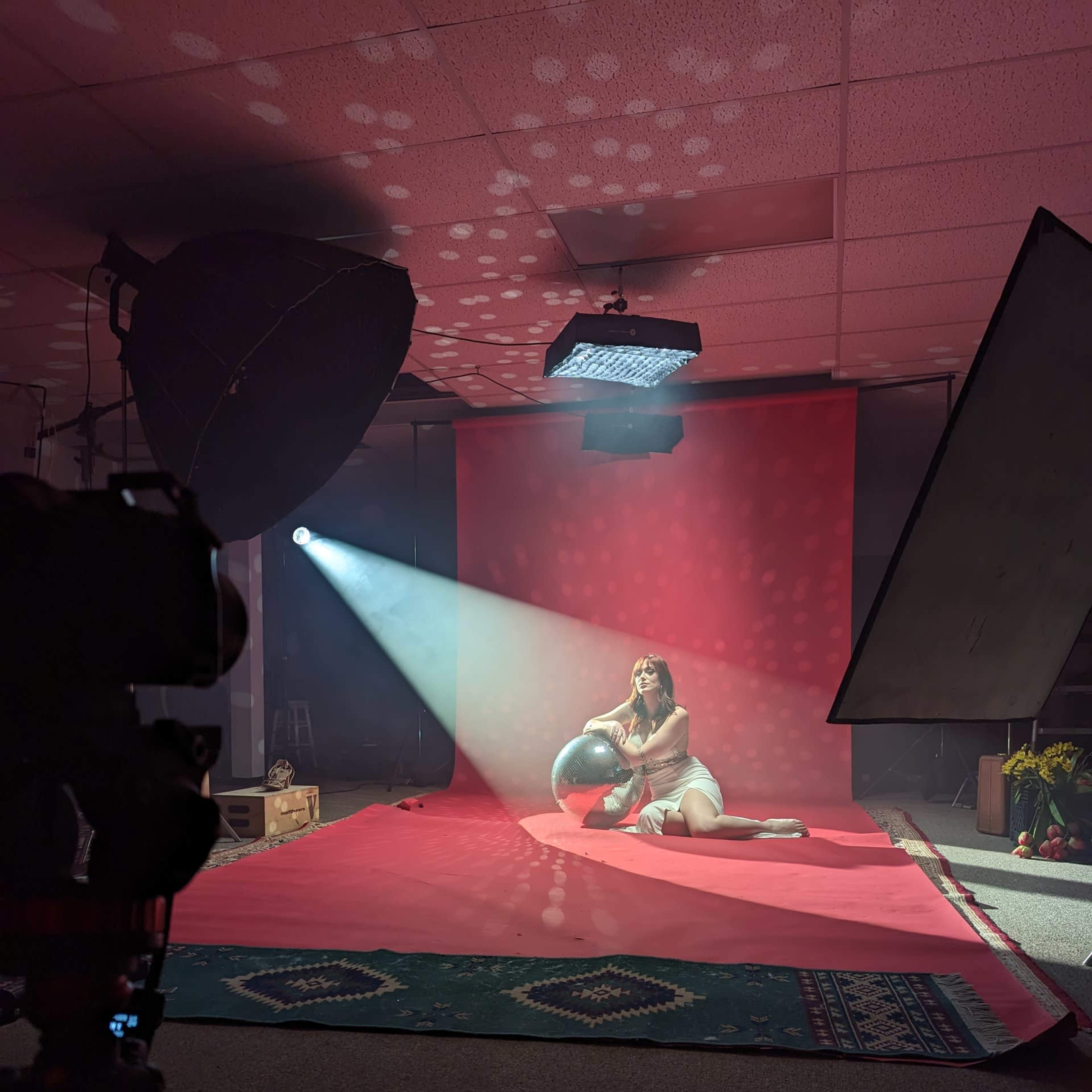A model sits on a red backdrop in a photography studio, holding a globe while light beams illuminate the scene.