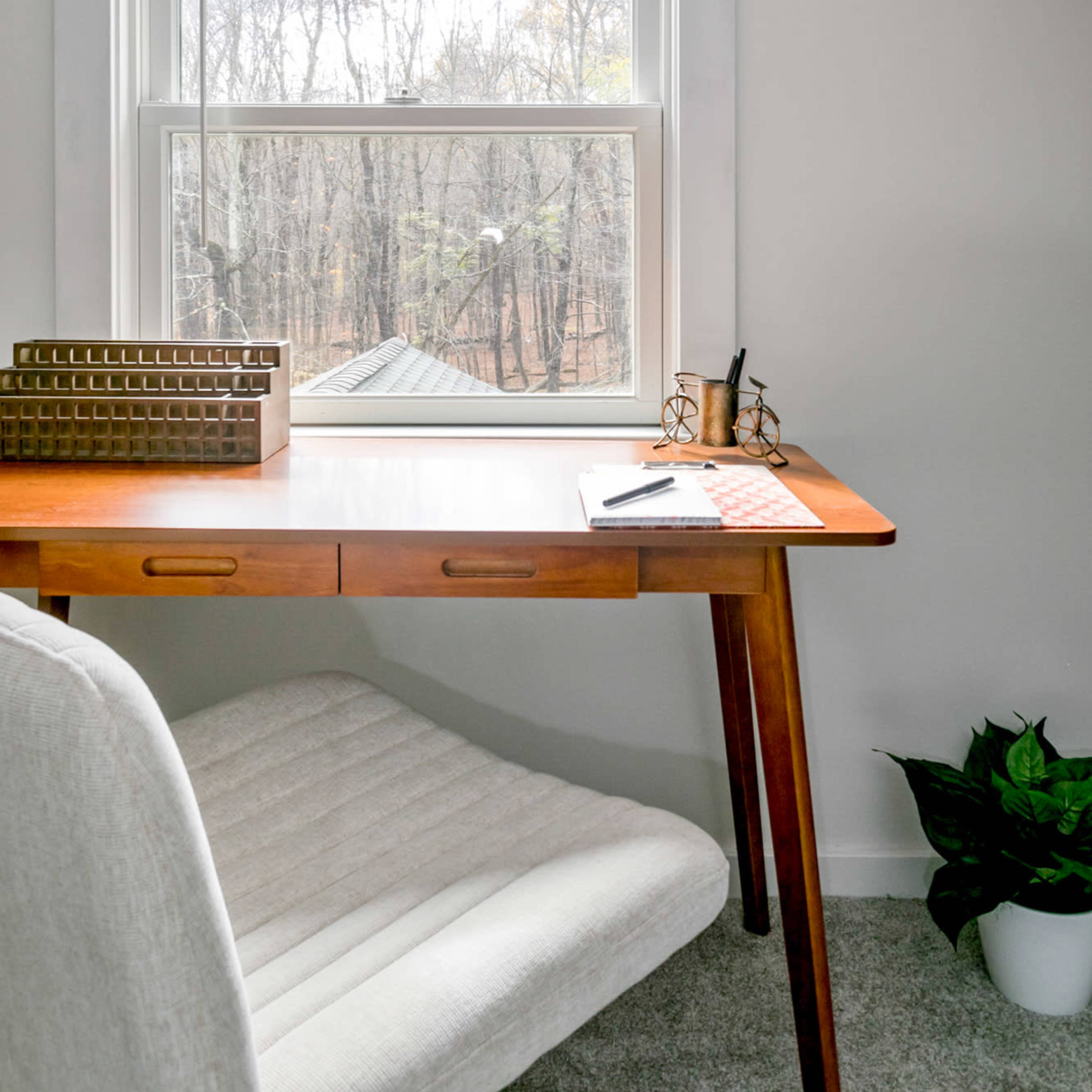 A wooden desk with a paper, pen, and a geometric decor piece sits by a window overlooking a wooded area.