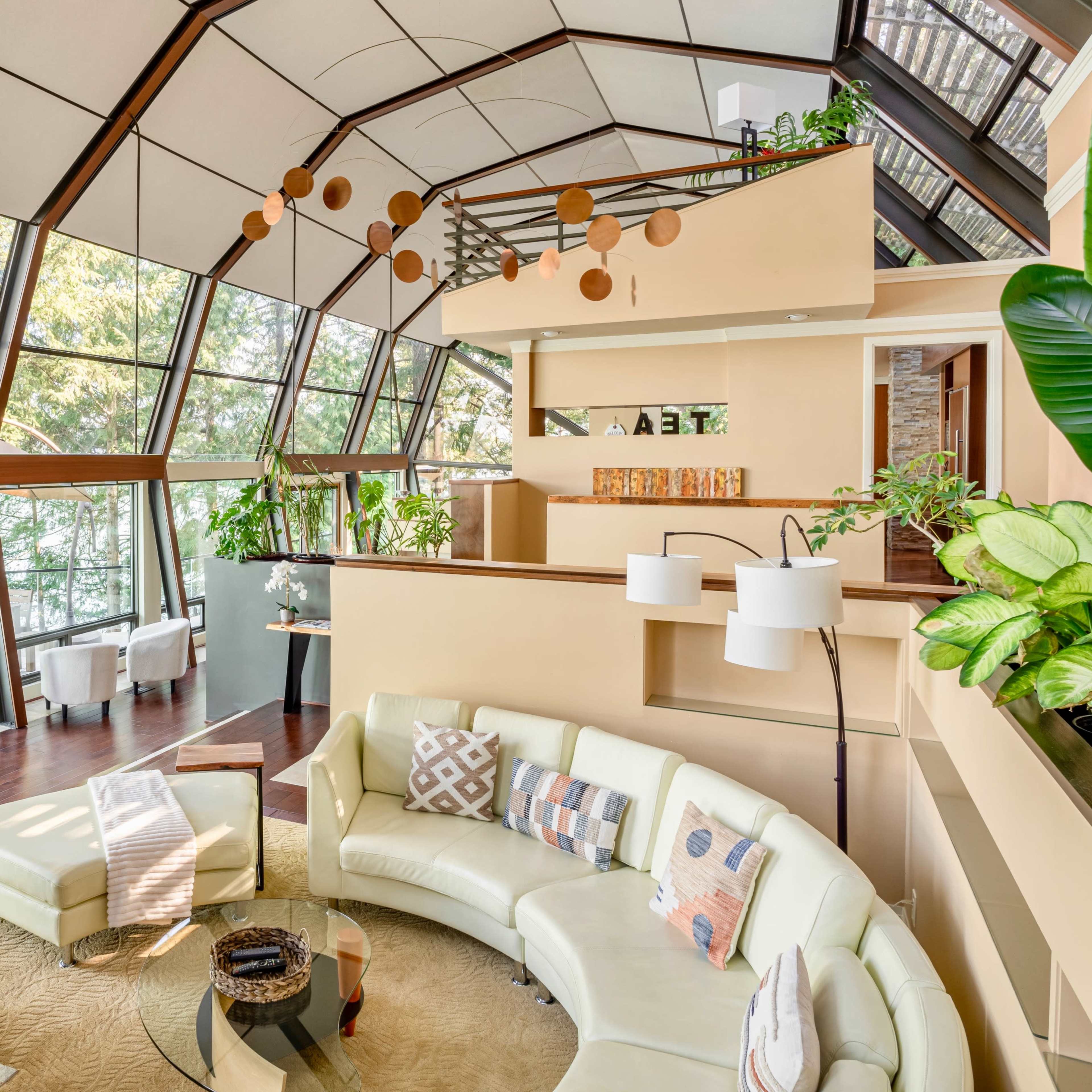 A spacious, modern living room with large windows, a curved white sofa, and various plants, creating a bright and airy atmosphere.