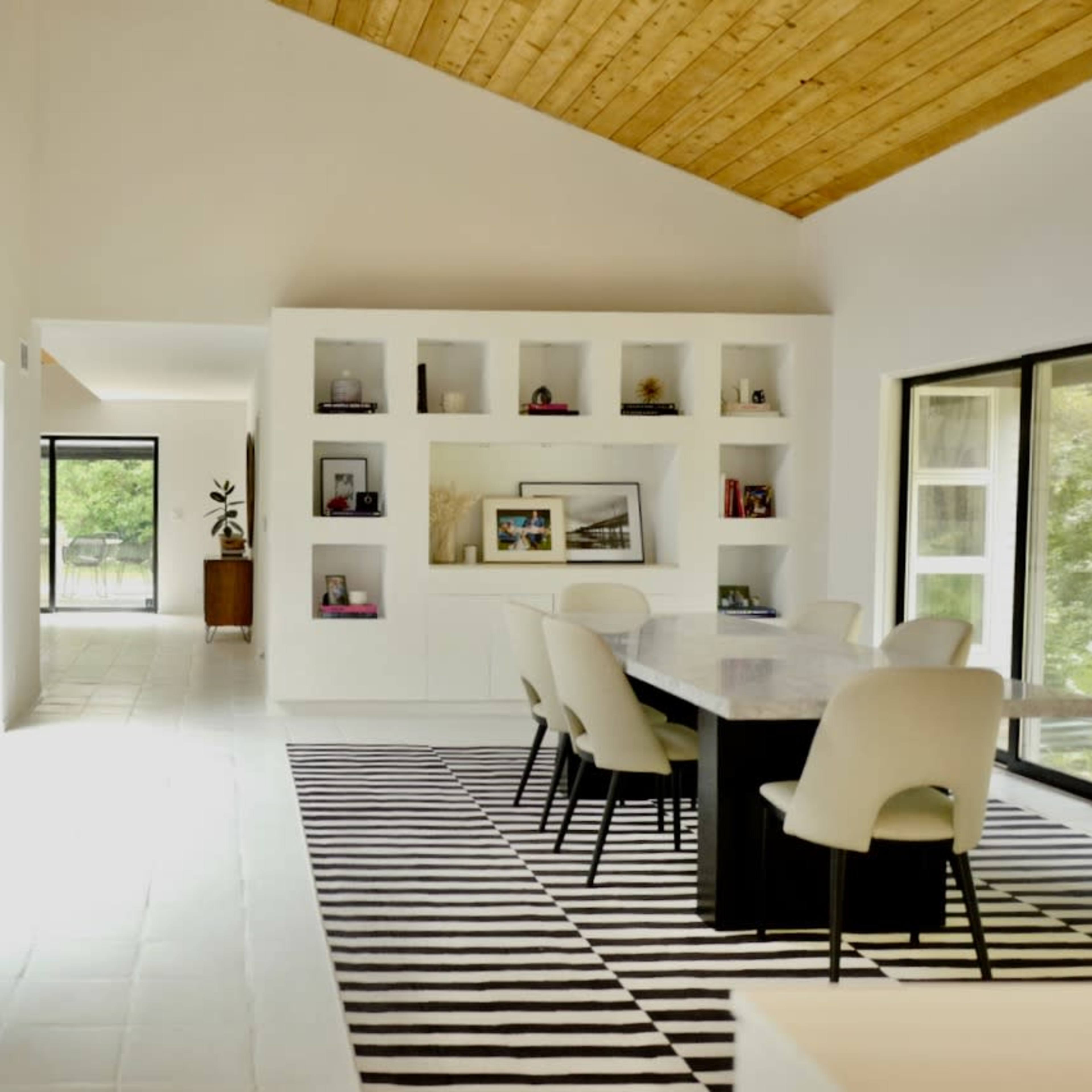 A spacious dining area with a large table surrounded by chairs, framed by large windows and a wall of shelves, featuring a modern wooden ceiling.