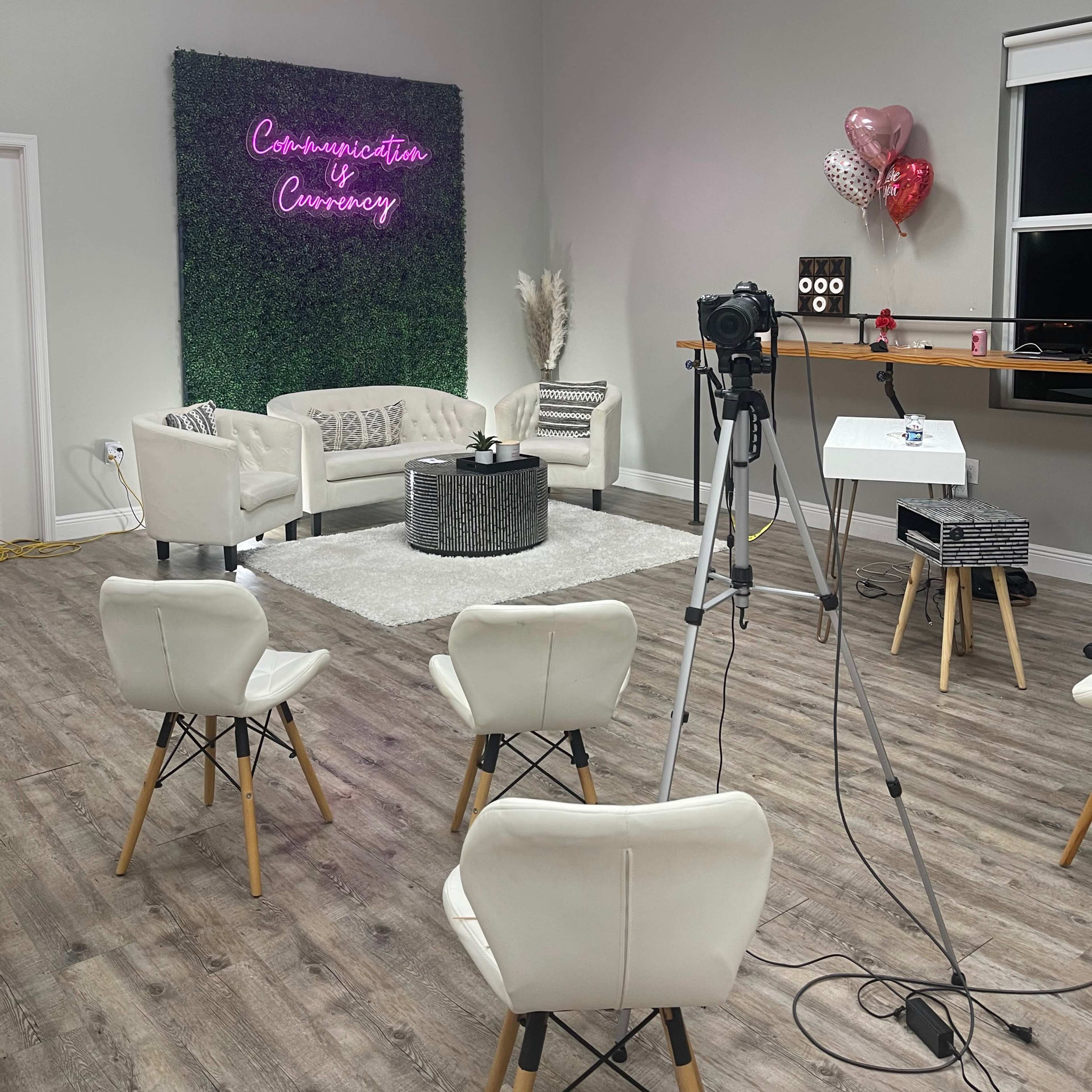 A cozy studio setup with a seating area, a neon sign on a green wall, and a camera pointed towards the seating arrangement.