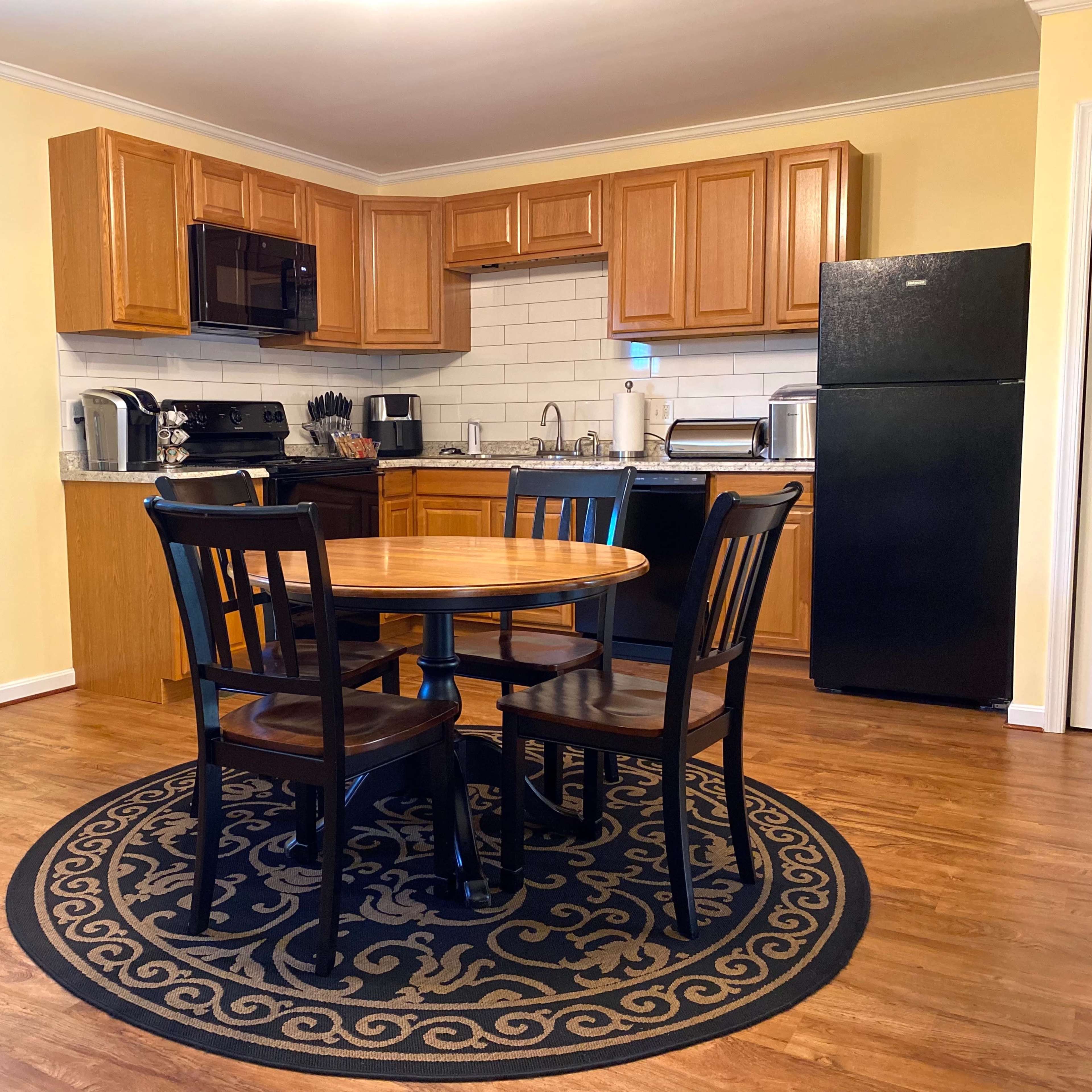A small dining area with a round wooden table surrounded by four black chairs, located beside a kitchen with wooden cabinets and a black refrigerator.