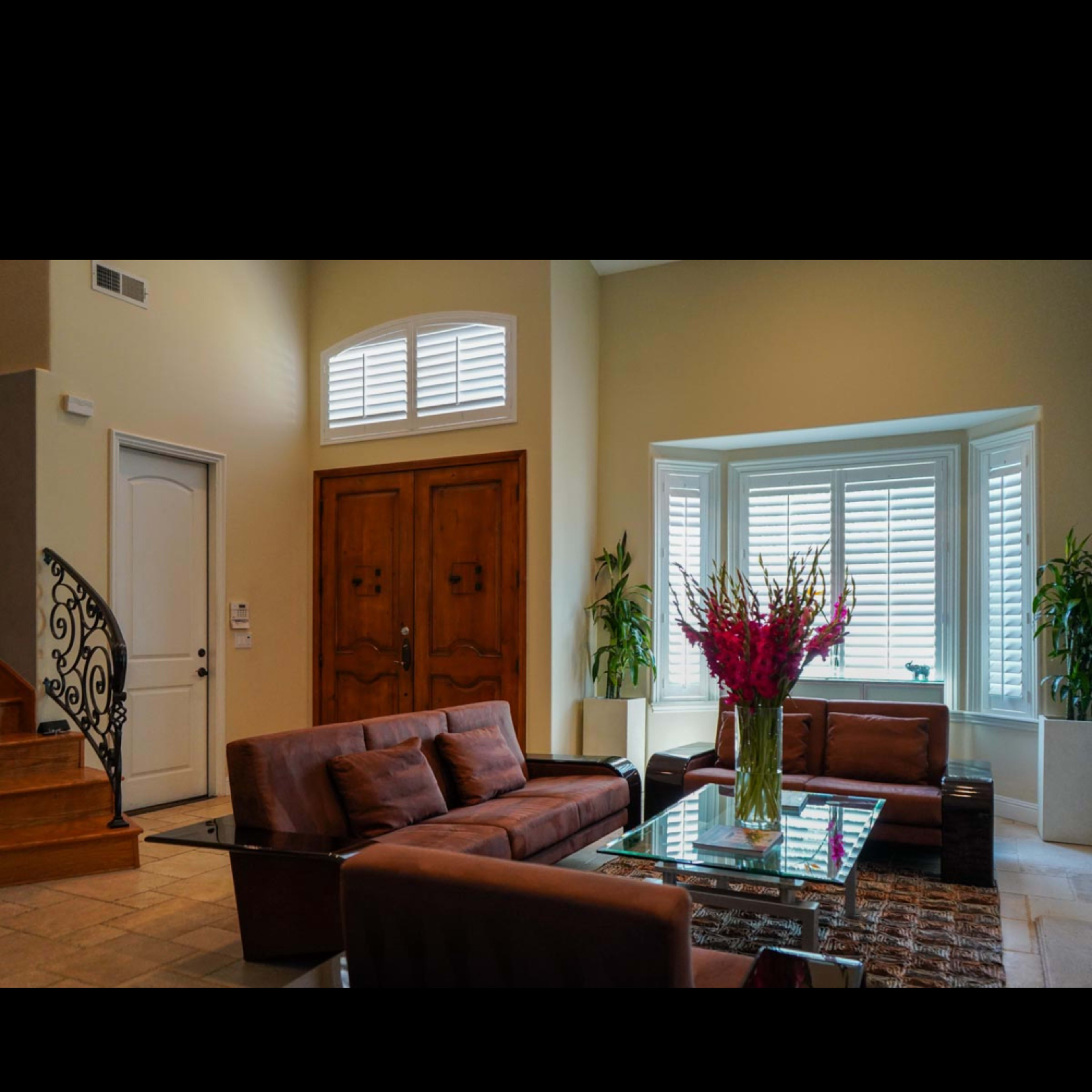 A modern living room features two brown sofas, a glass coffee table, and large windows with plants nearby, set against a backdrop of a staircase and decorative doors.