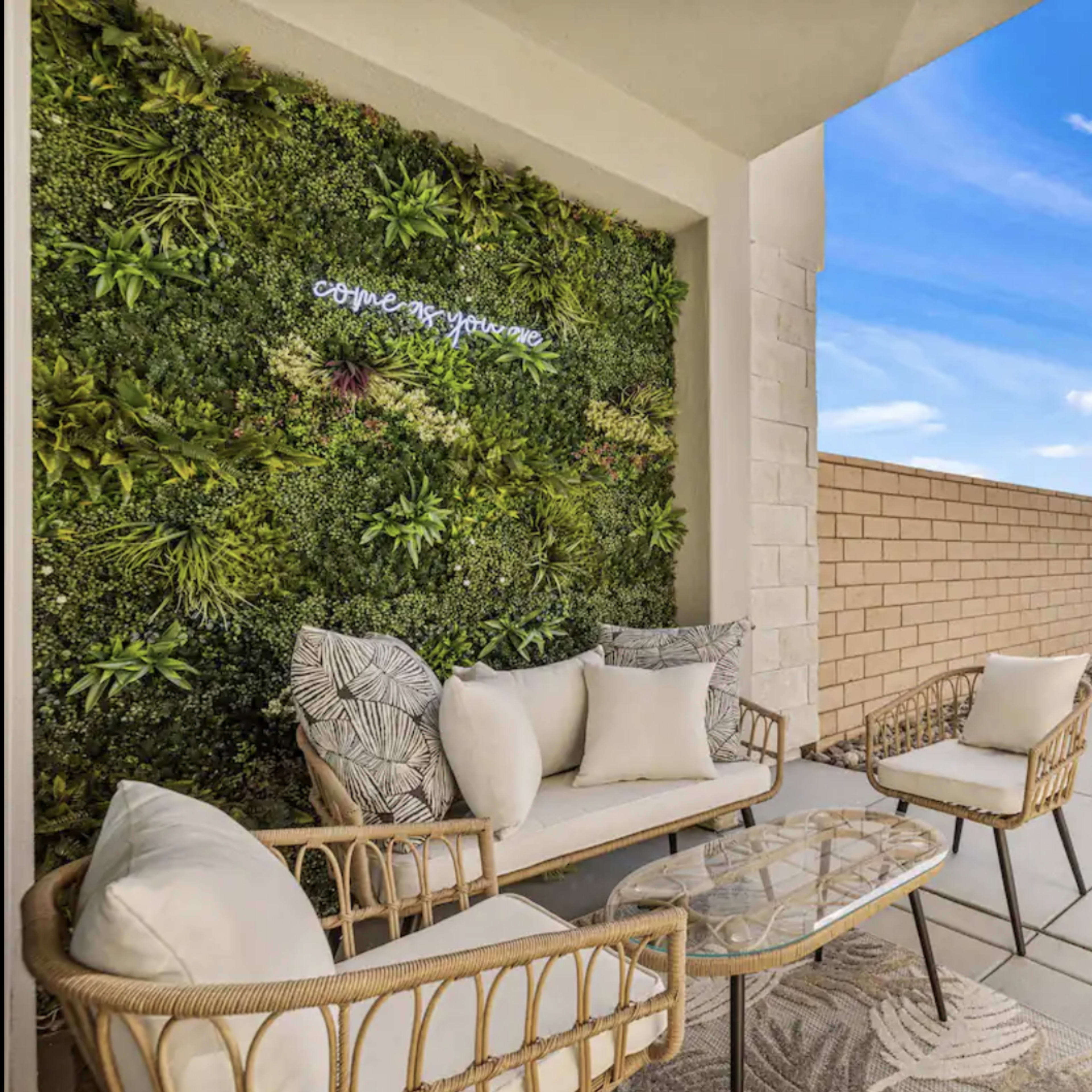 A seating area features two wicker chairs and a glass table, framed by a vertical garden and overlooking a body of water and a distant house.