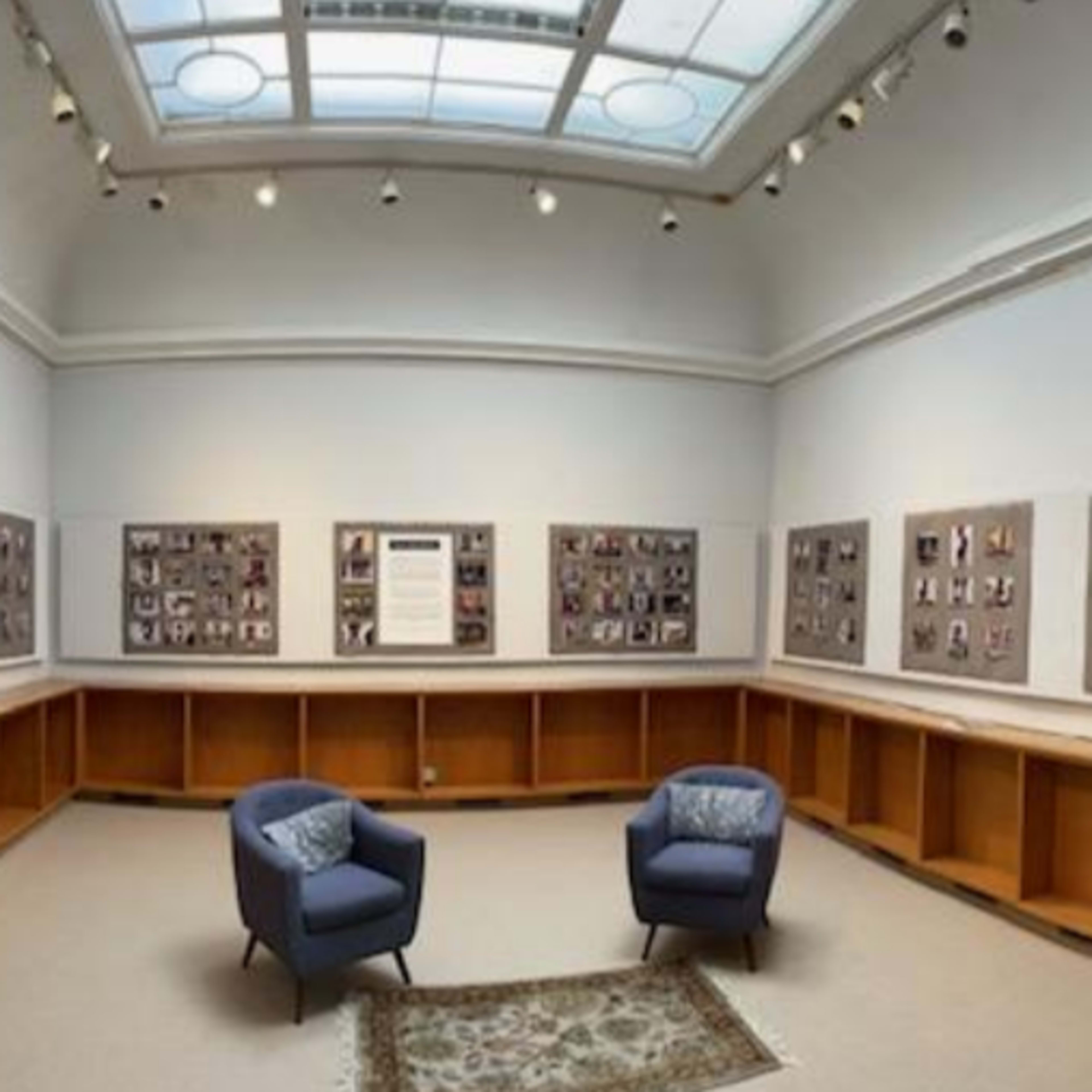A gallery room with two blue chairs situated on a carpet, surrounded by walls displaying framed photographs.