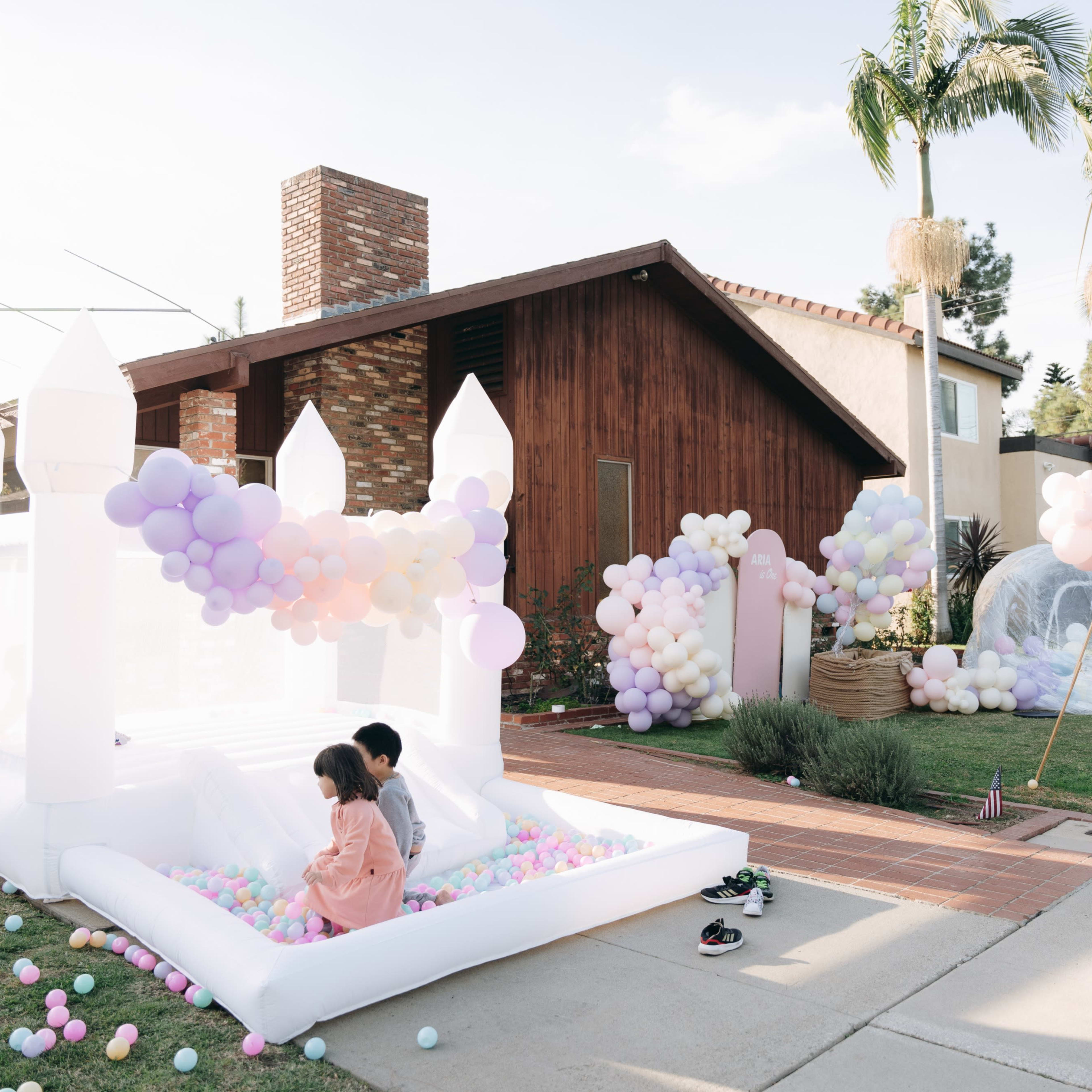 A bounce house filled with colorful balls is set up on a lawn in front of a house adorned with balloon decorations.