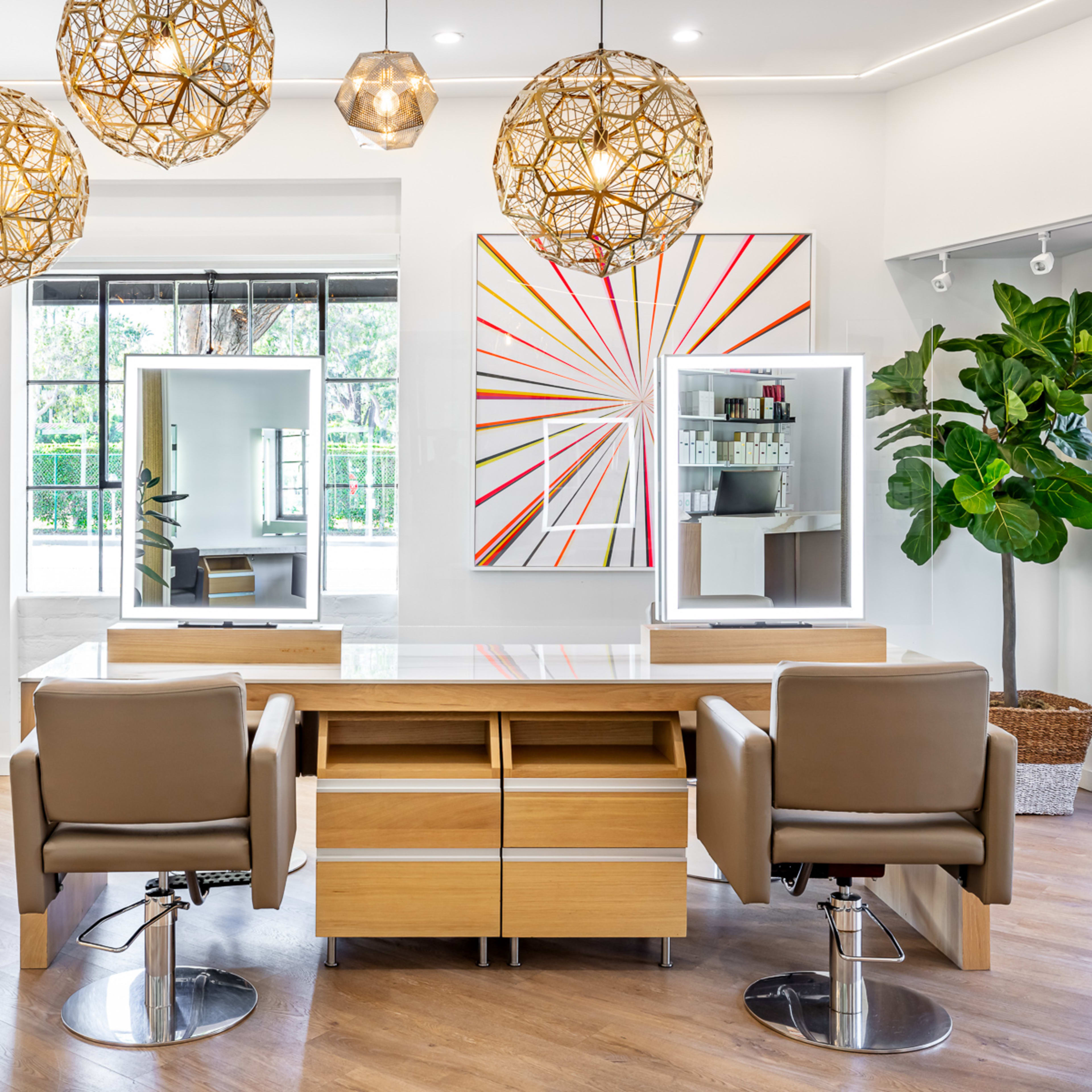 A modern salon interior featuring two styling stations with mirrors, wooden furniture, decorative hanging lights, and large windows with views of greenery.