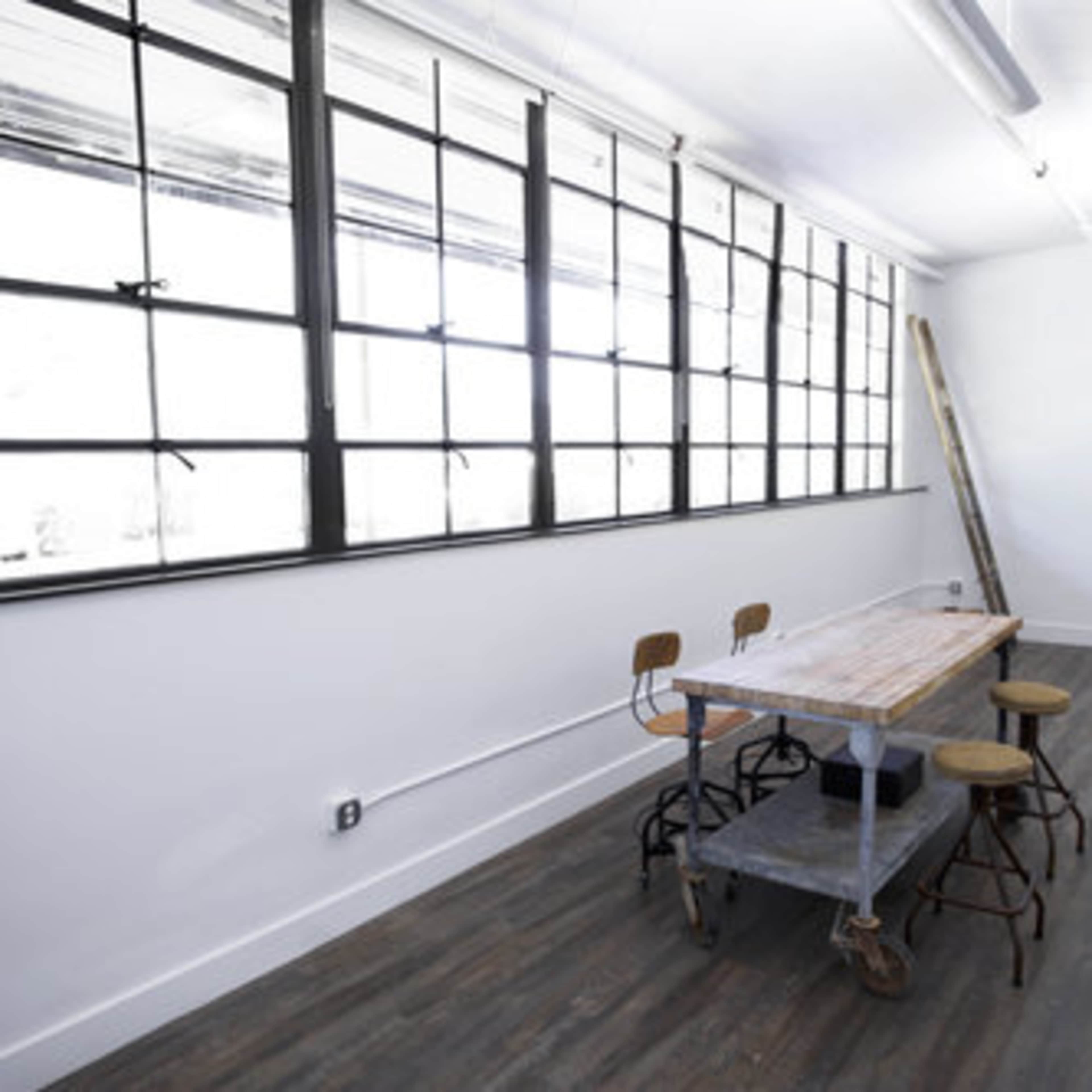 A minimalist room with large windows, a wooden table on wheels, and four metal chairs arranged around it.