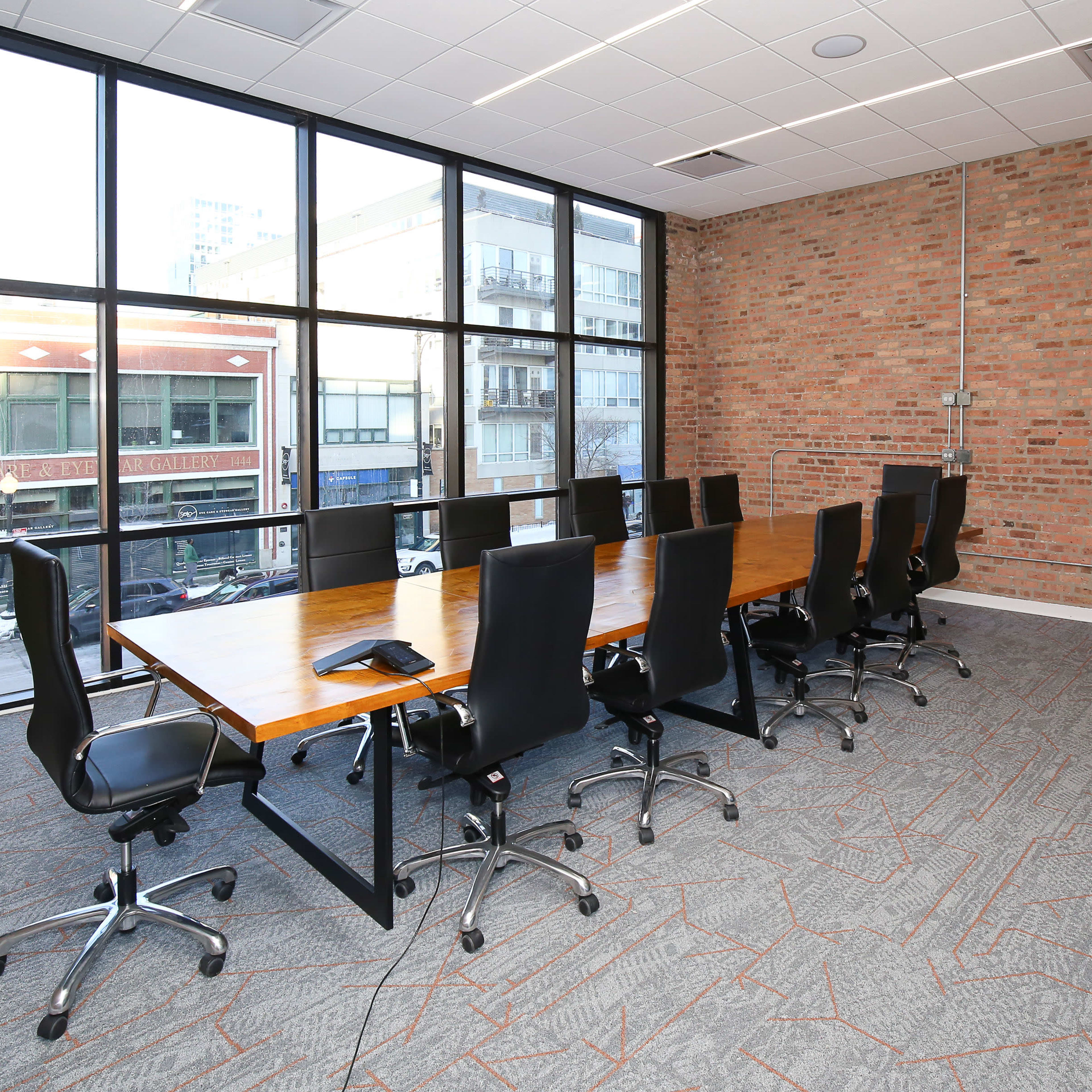 A modern conference room features a large wooden table surrounded by black rolling chairs, with large windows providing a view of the outside.