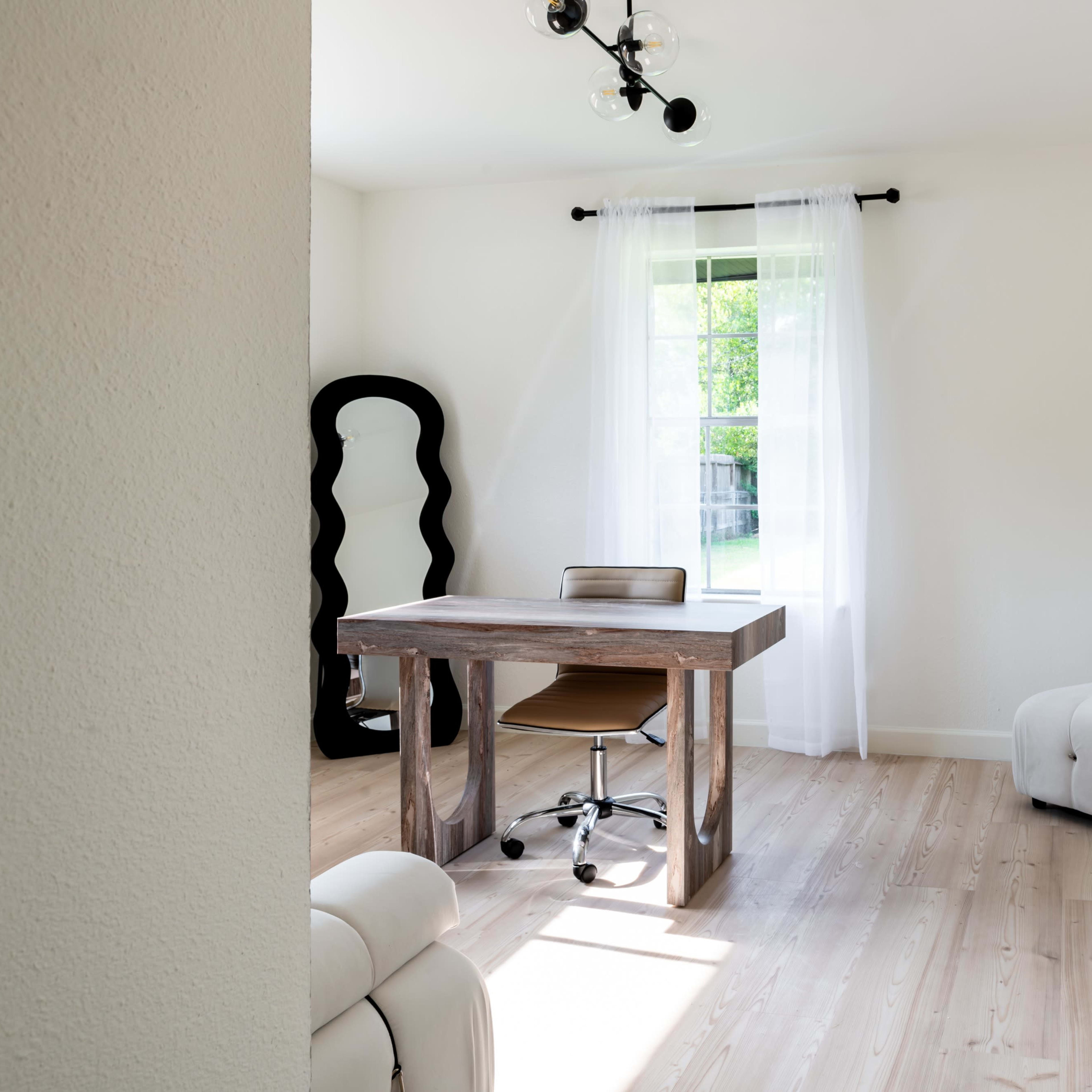 A minimalist home office features a wooden desk, a chair, and a light-colored couch, with natural light illuminating the space through a window.