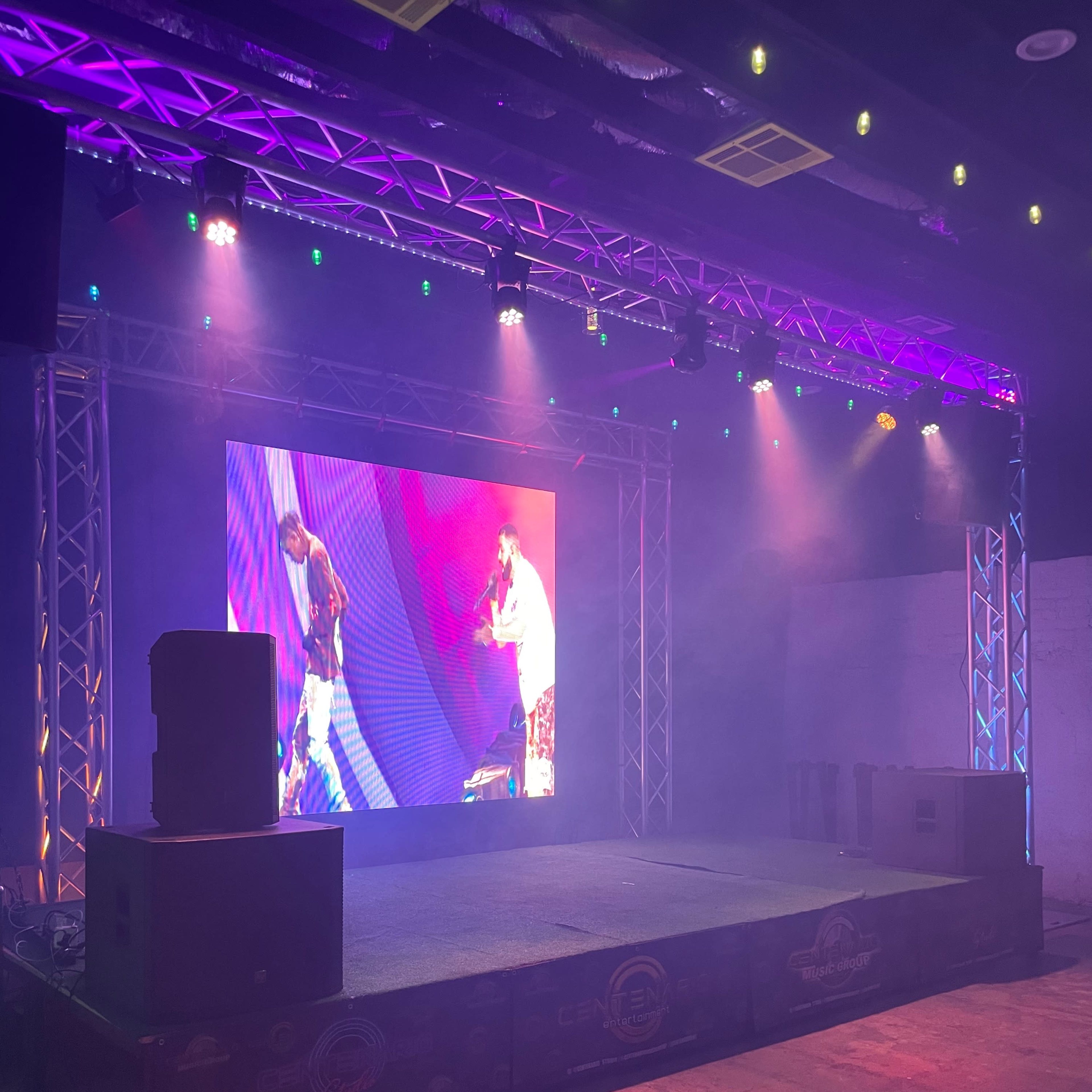 A stage setup features a large screen and lighting effects, surrounded by sound equipment, in a dimly lit venue.