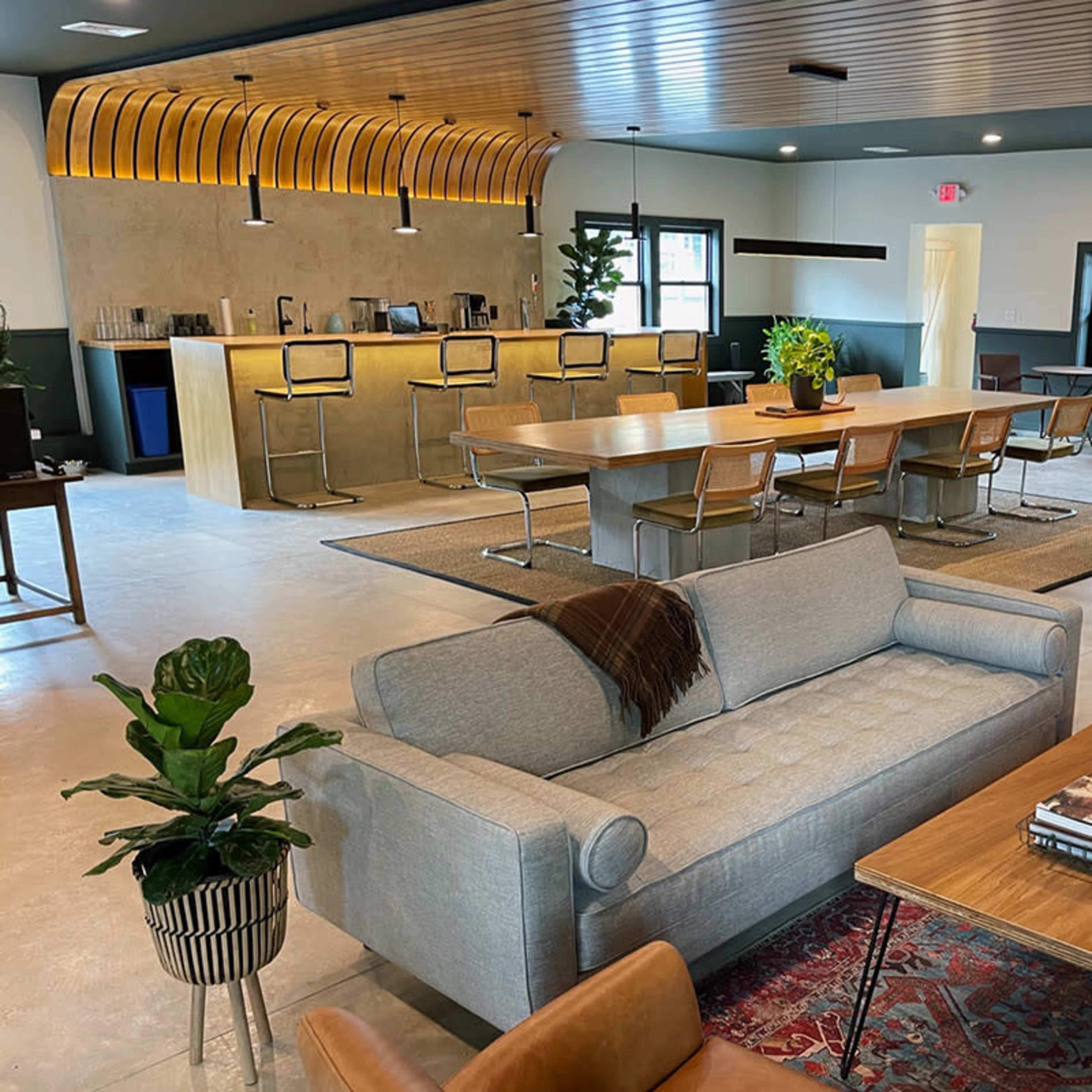 A modern interior space featuring a communal seating area with a sofa and armchairs, a large wooden dining table, and a bar counter in the background.