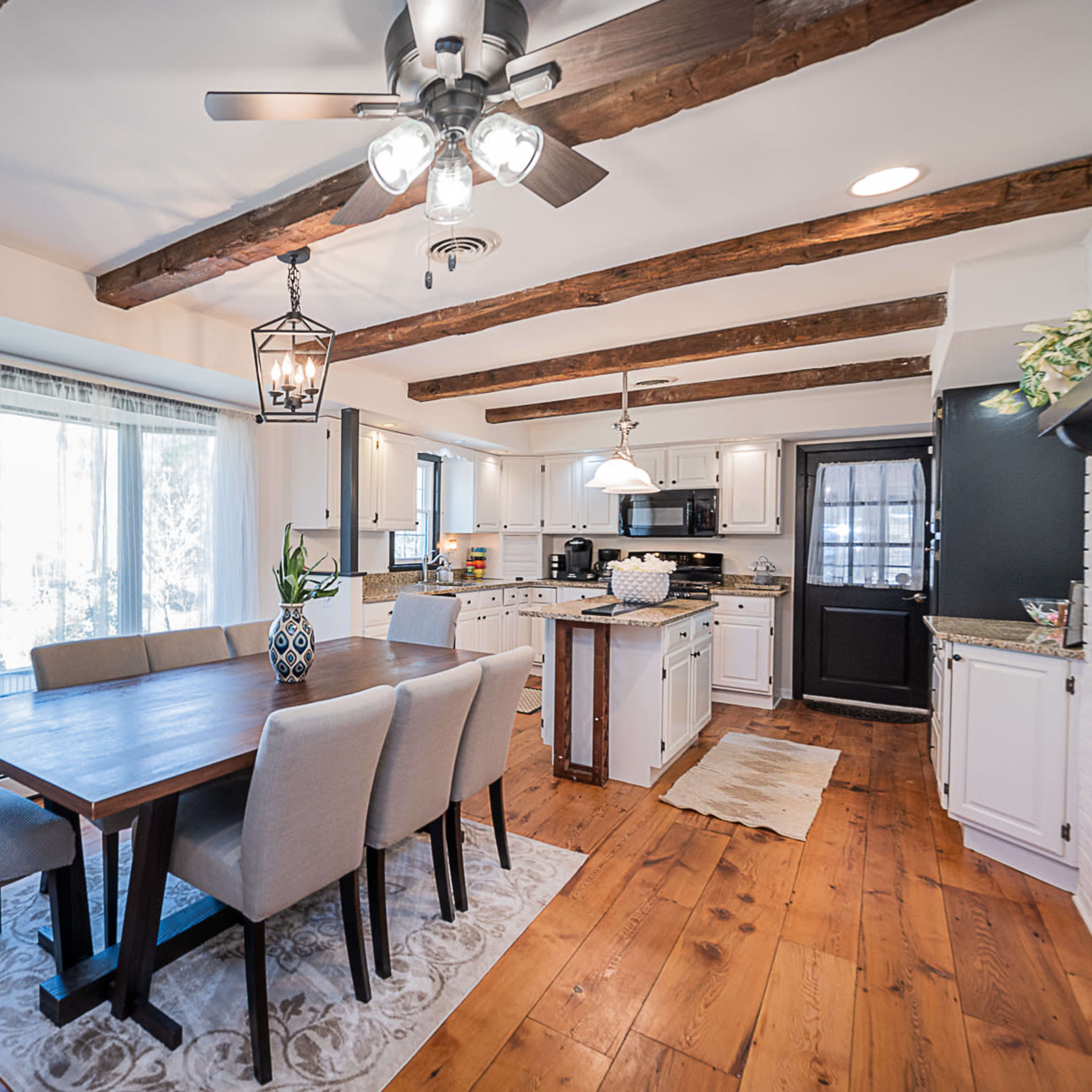 A bright kitchen features wooden beam ceilings, a dining table with upholstered chairs, and modern appliances.