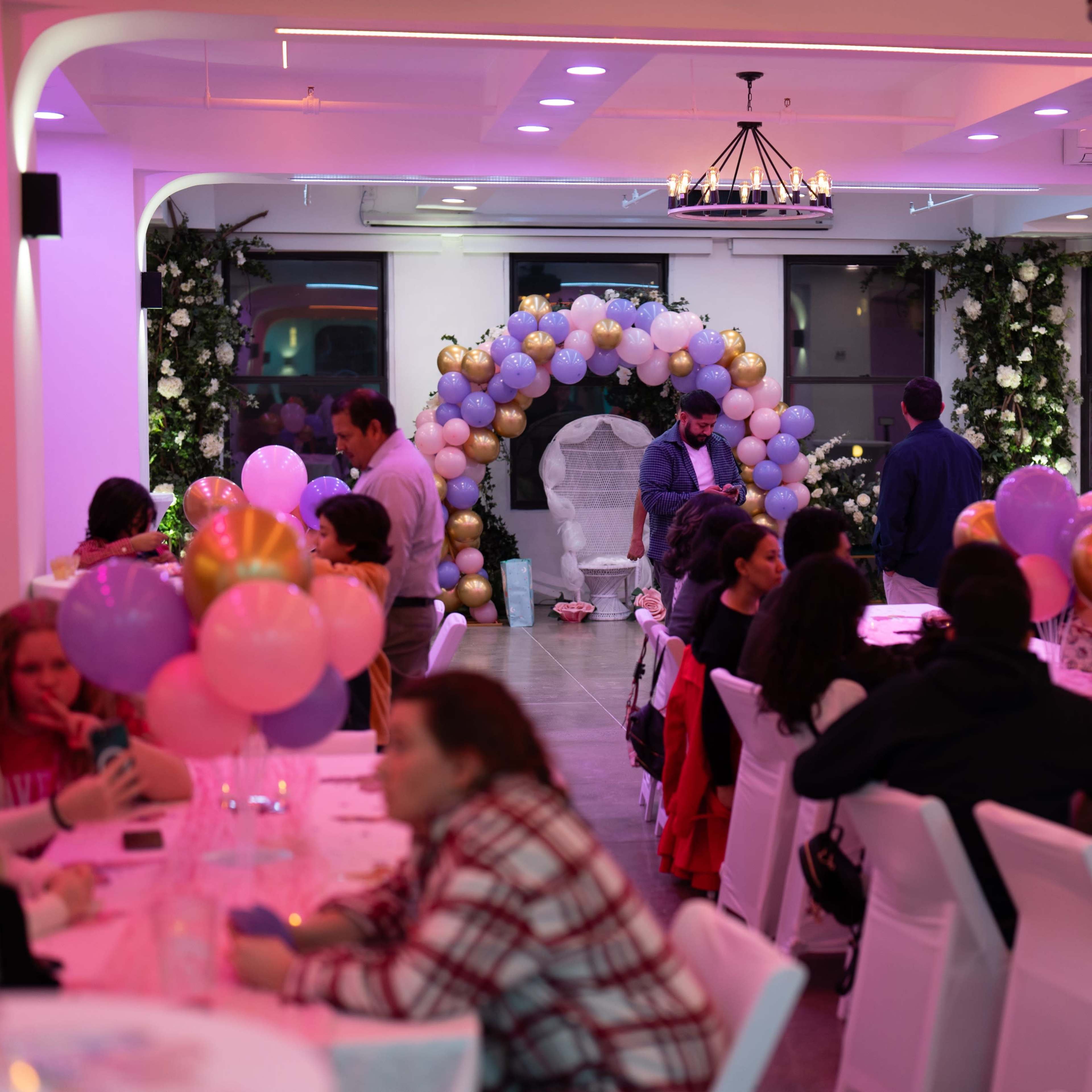 A decorated event space features tables with balloons and a floral arch in the background, with guests seated and socializing.