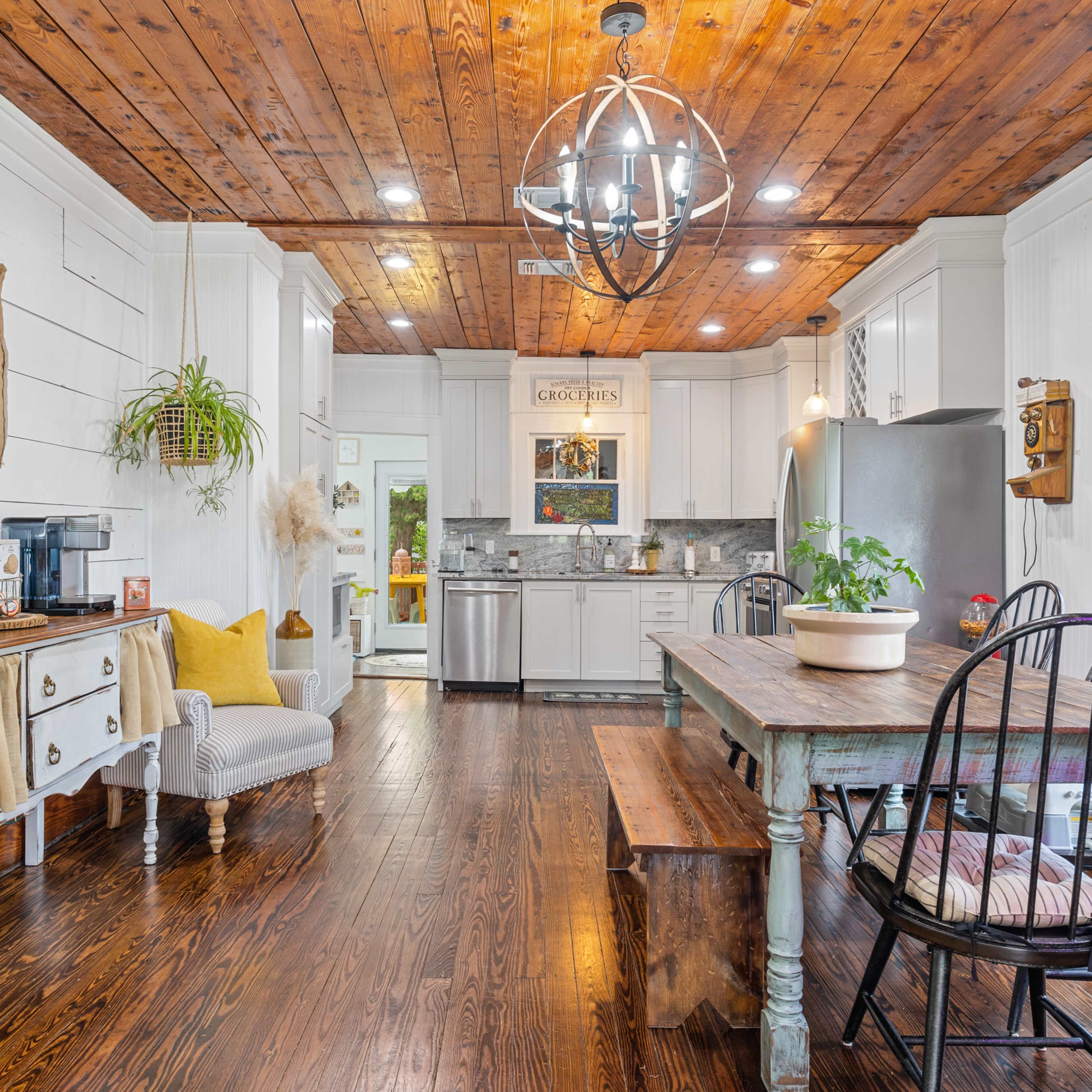 A well-lit kitchen with a wooden ceiling, a large dining table, and a coffee station beside a comfortable seating area.