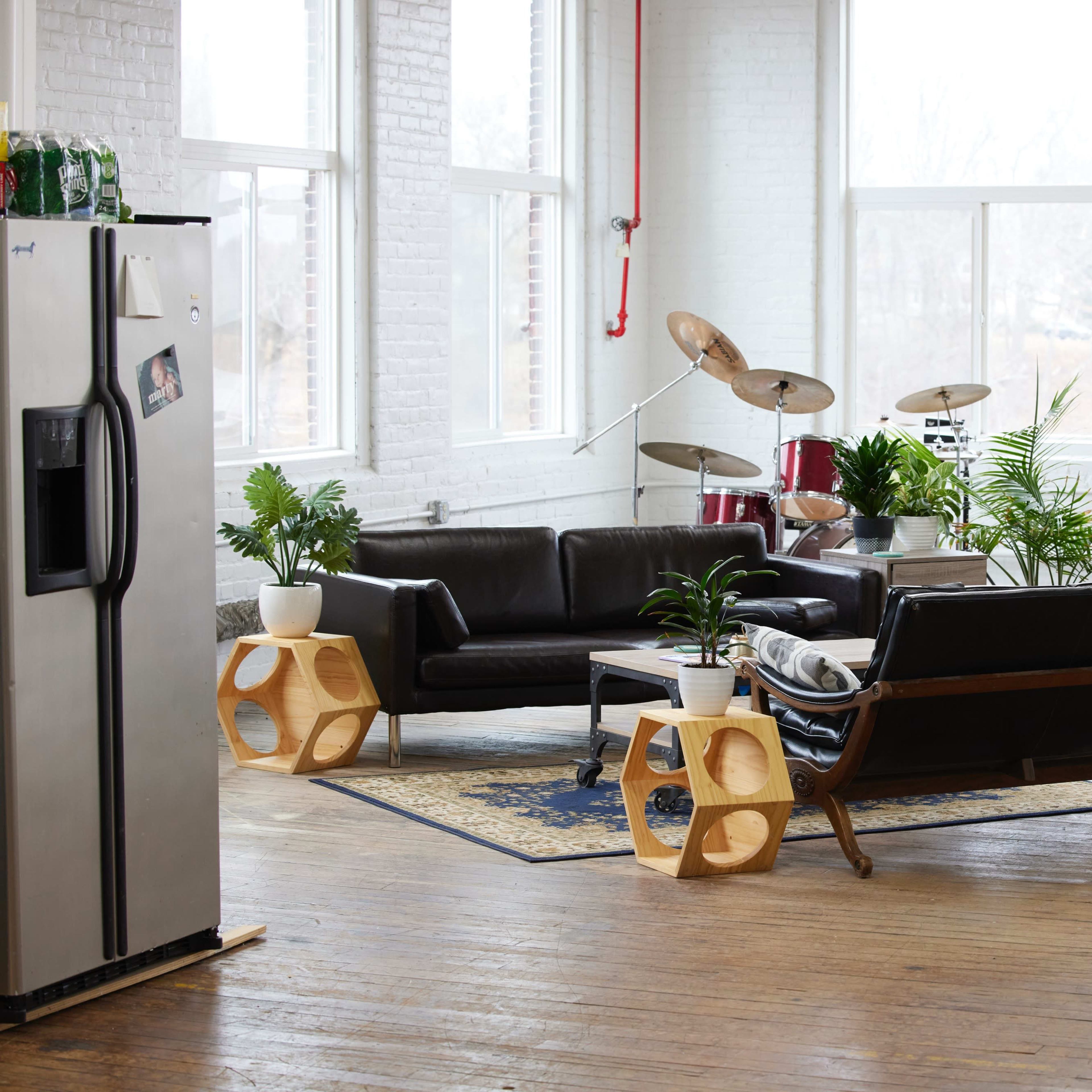 A spacious, well-lit room with two black sofas, a coffee table, a few potted plants, a modern-style rug, and a drum set in the corner.