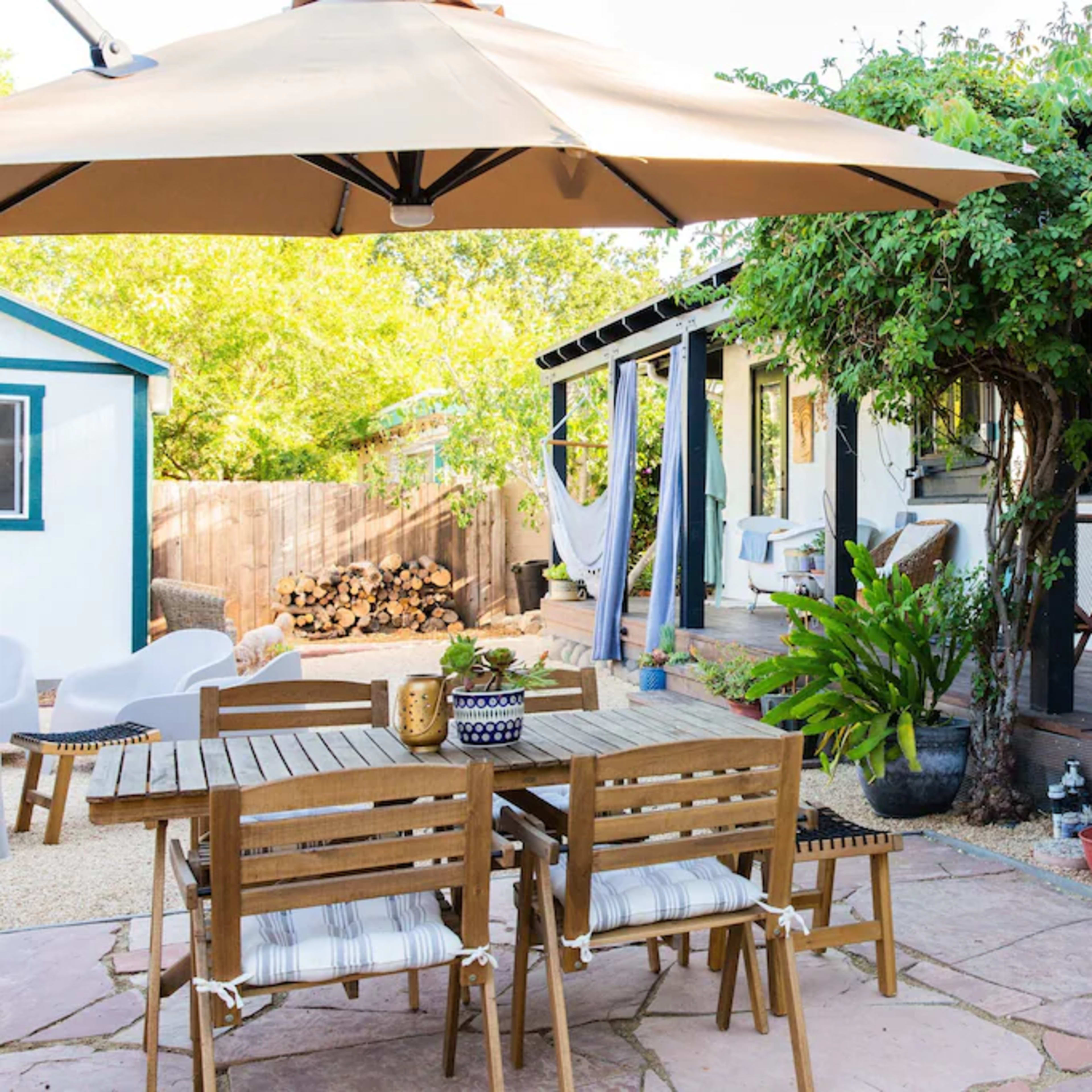 A patio area features a wooden dining table with chairs, an umbrella above, and a well-kept garden leading to a house with a porch.