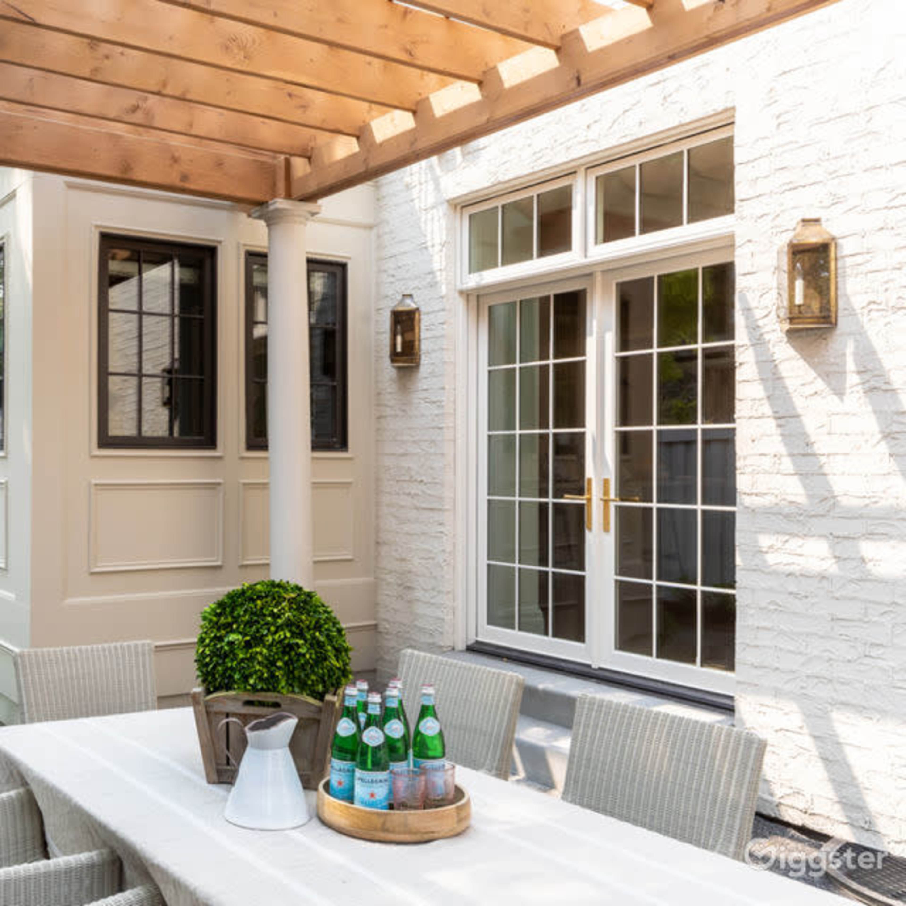 A patio area features a dining table surrounded by chairs under a wooden pergola, with a pathway leading to a gated entrance filled with greenery.