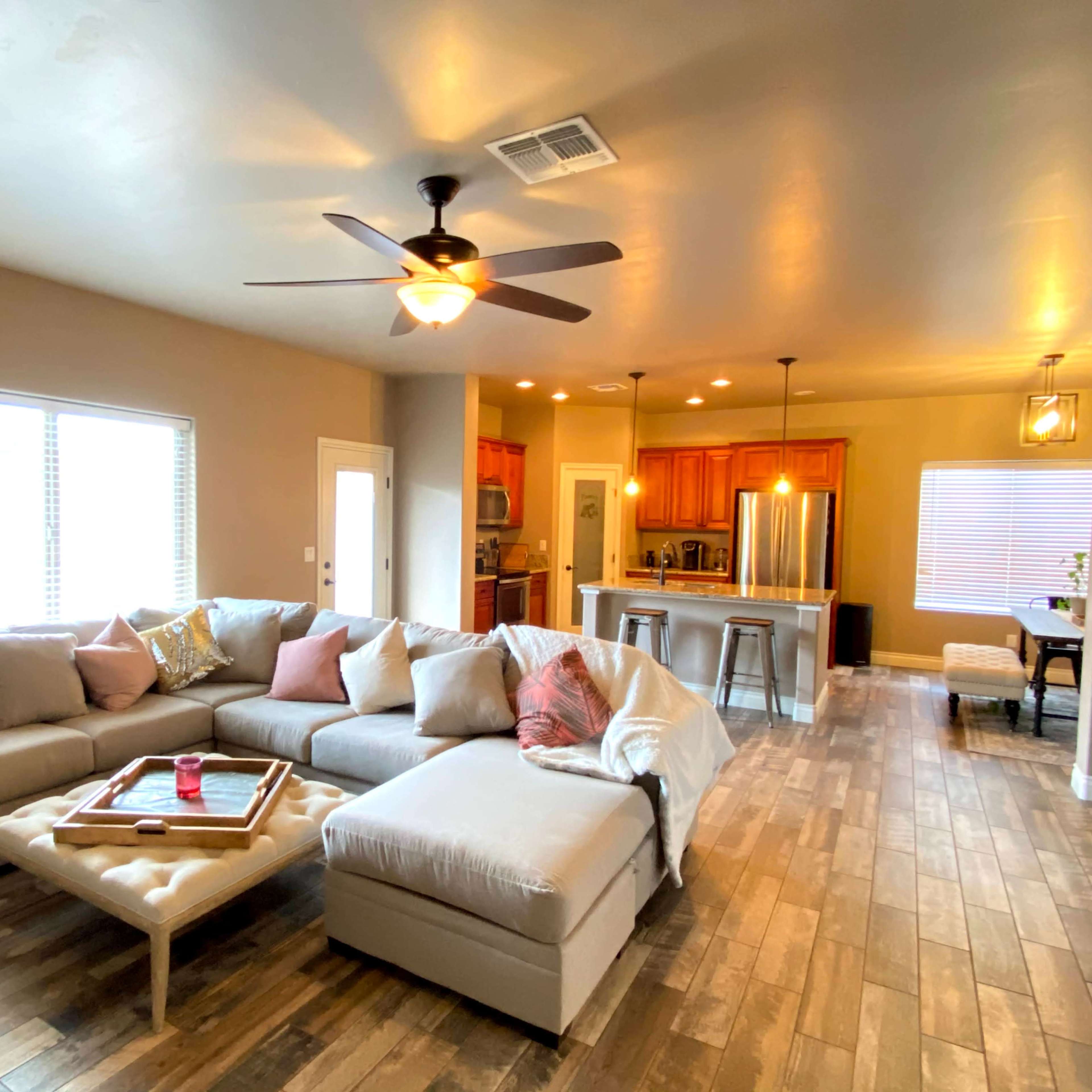 A spacious living room features a large sectional sofa, a coffee table, and an open kitchen area in the background with wooden cabinetry.