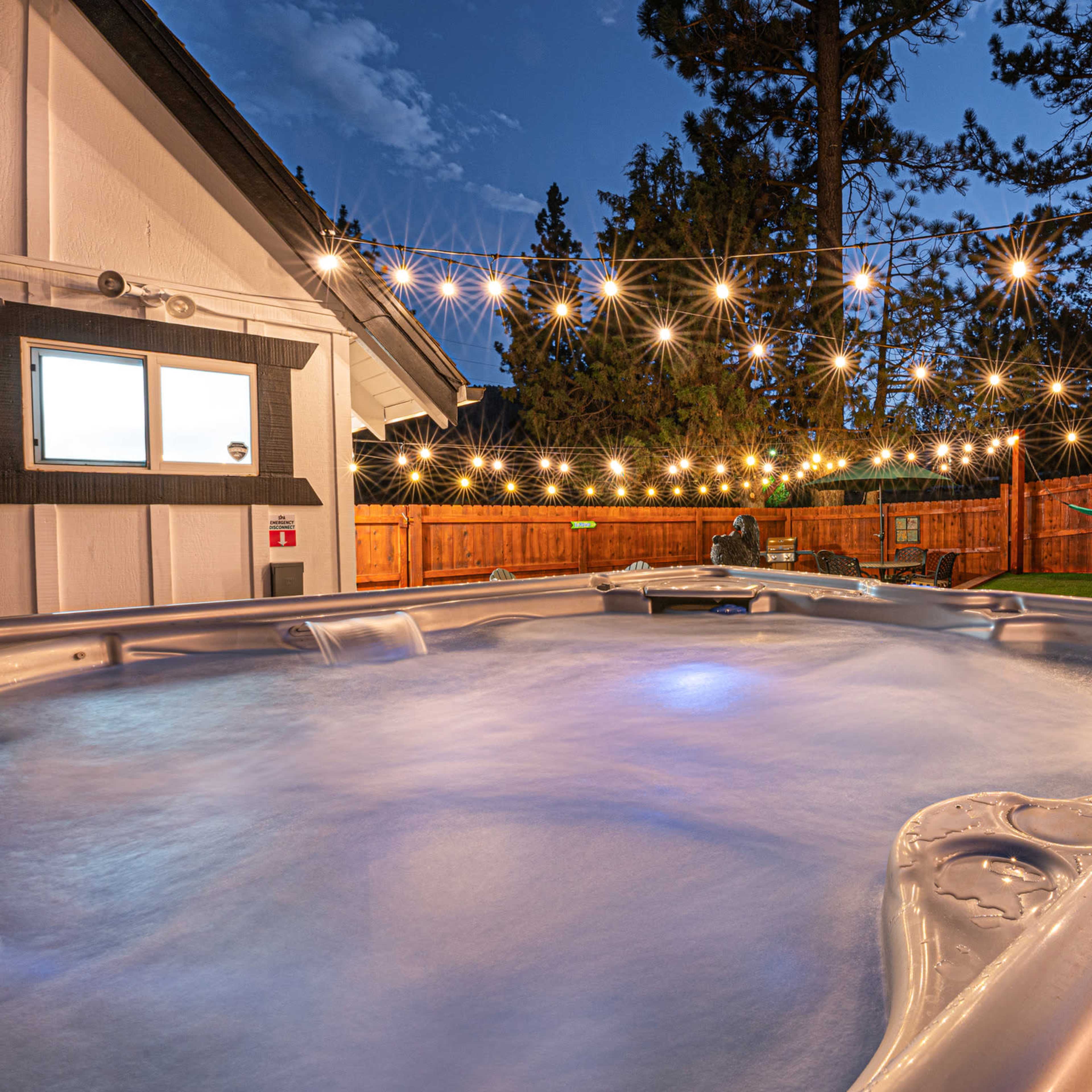 A hot tub with illuminated water sits in a backyard surrounded by string lights and a wooden fence, with a hammock and seating area in the background.