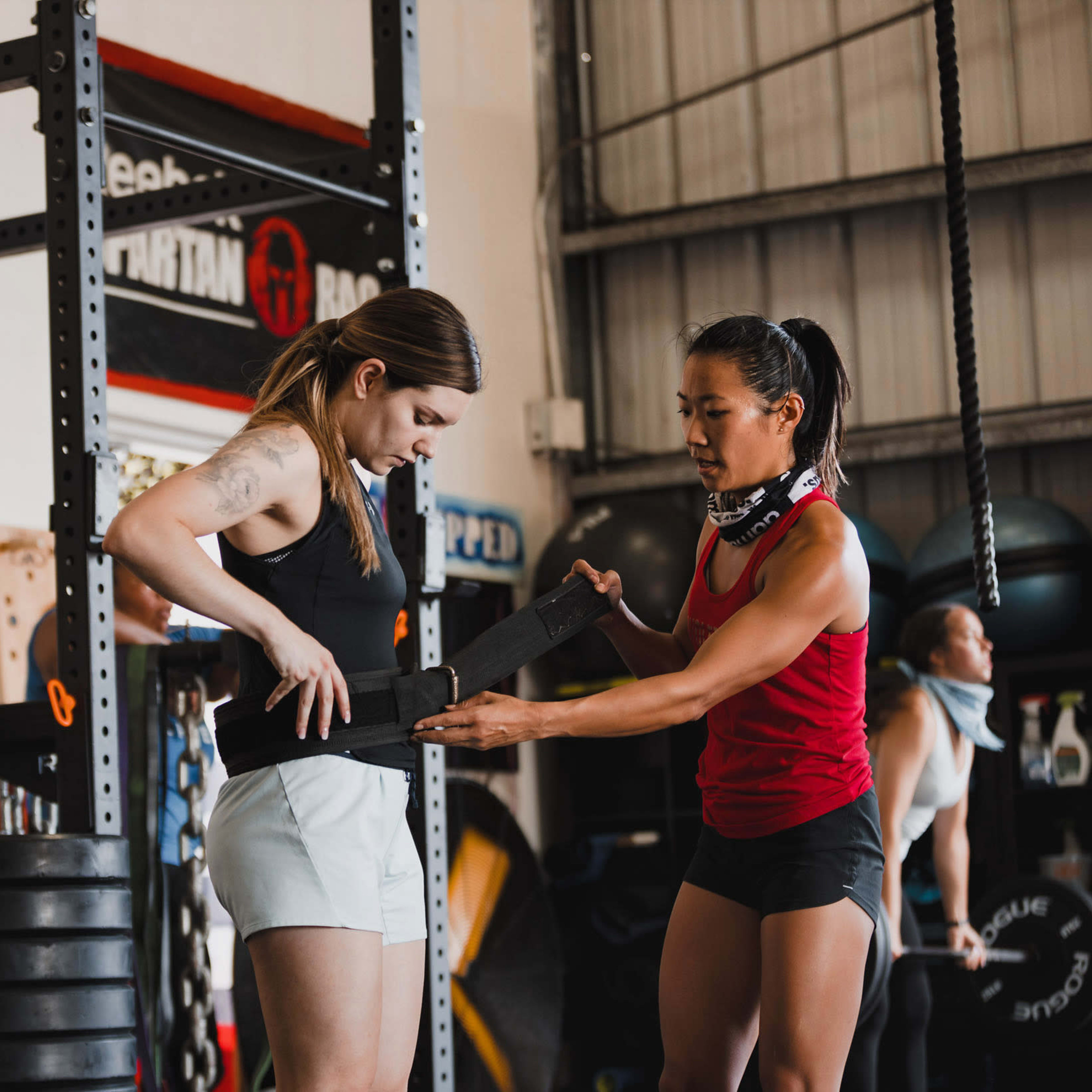 Two women are in a gym setting, with one adjusting a weightlifting belt on the other while exercise equipment is visible in the background.