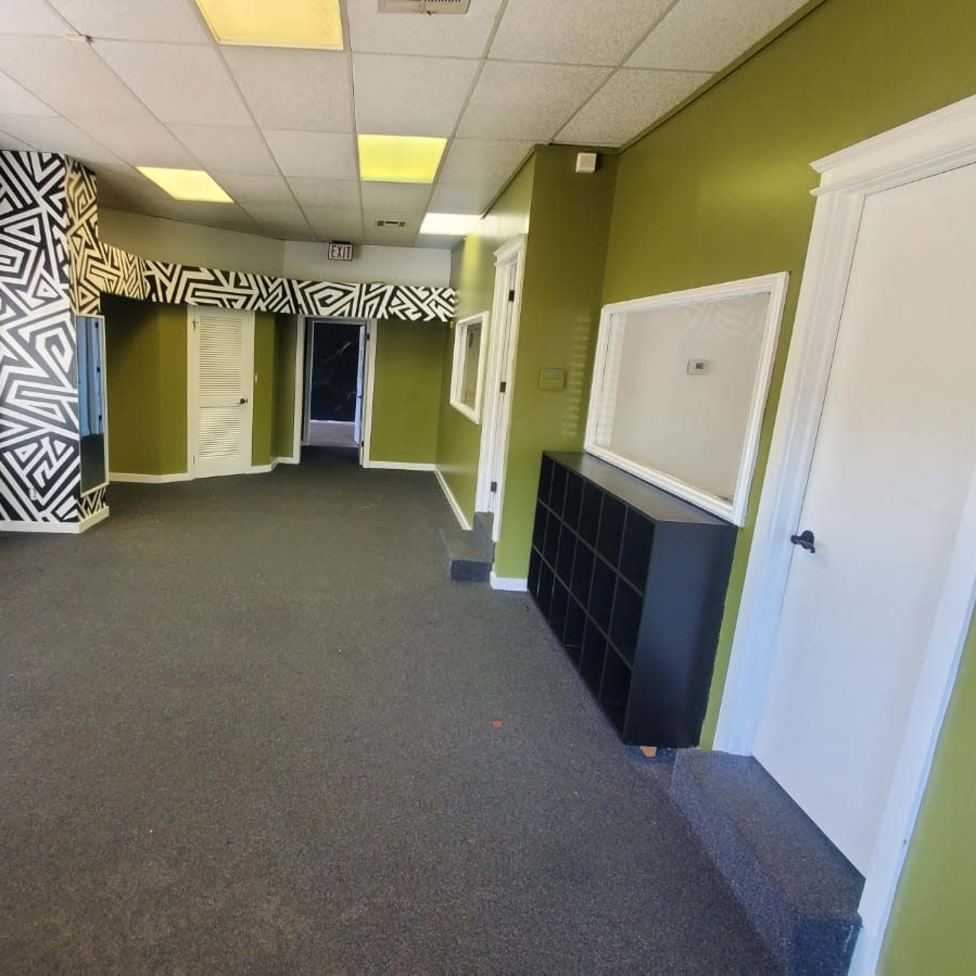 A spacious, brightly lit room with green walls and black-and-white patterned accents, featuring a gray carpet and several doorways.