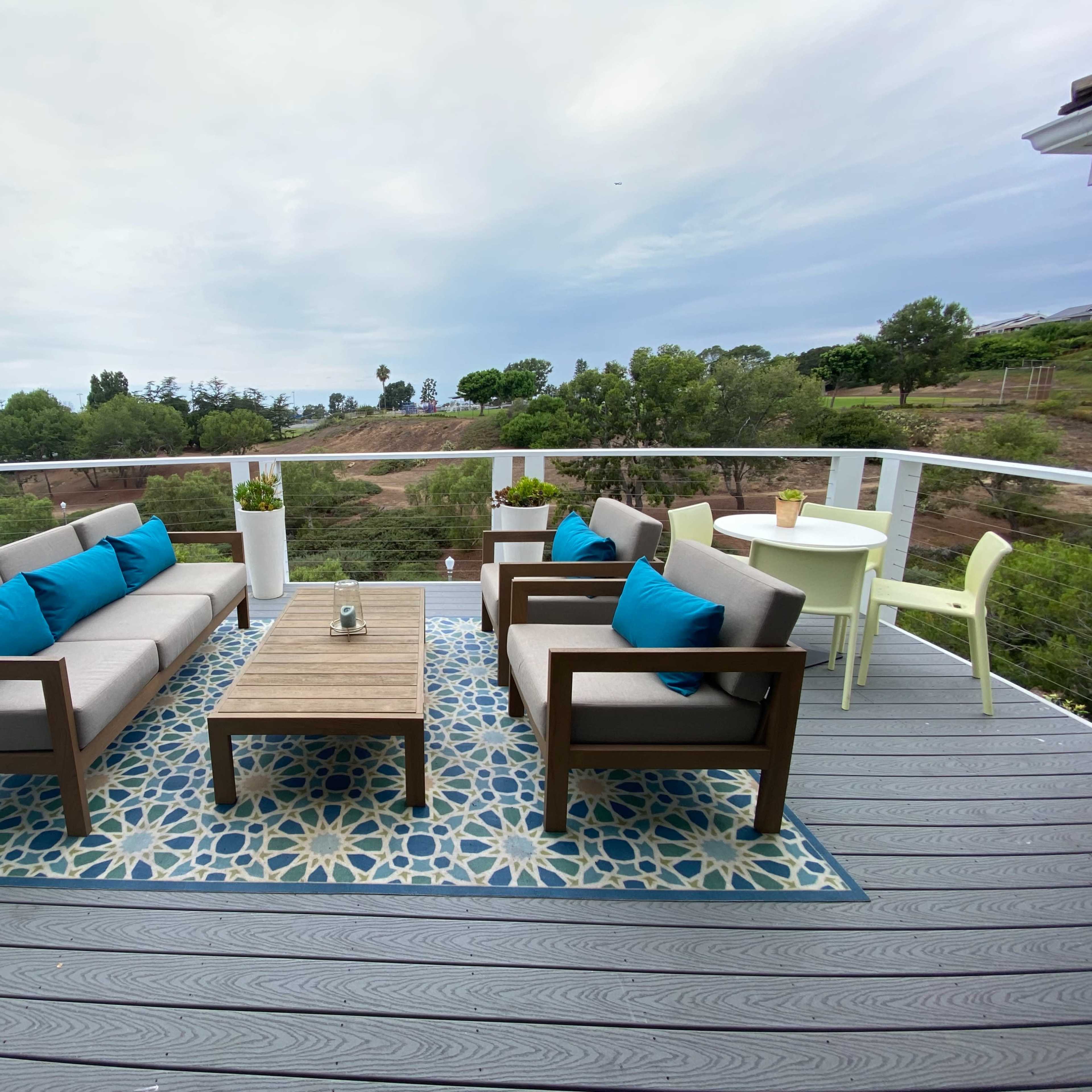 A modern outdoor patio features a seating area with two sofas and a coffee table, surrounded by a decorative rug, showcasing a view of lush greenery.