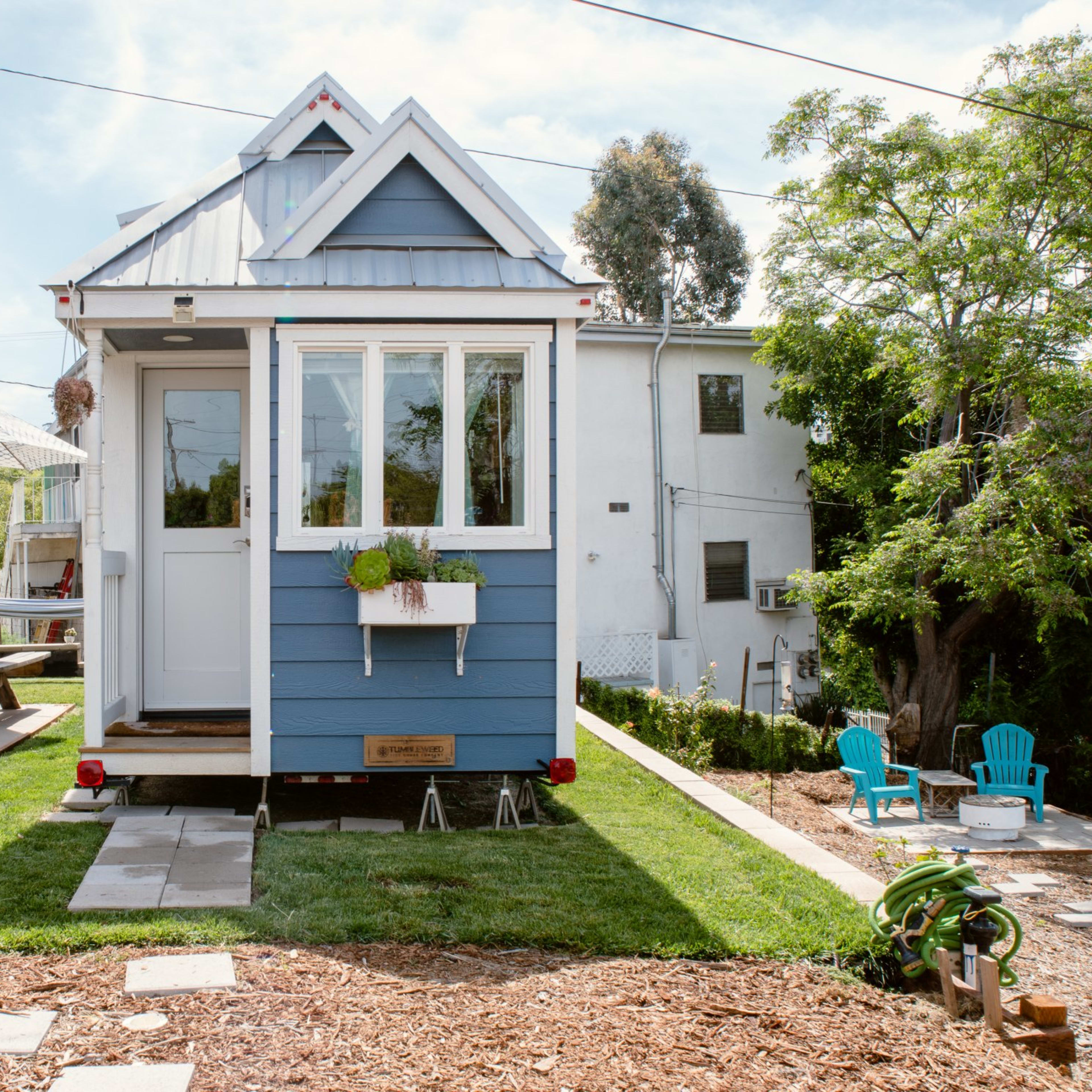 A small blue house with a peaked roof is set in a grassy yard, featuring a picnic table, barbecue grill, and bicycles nearby.