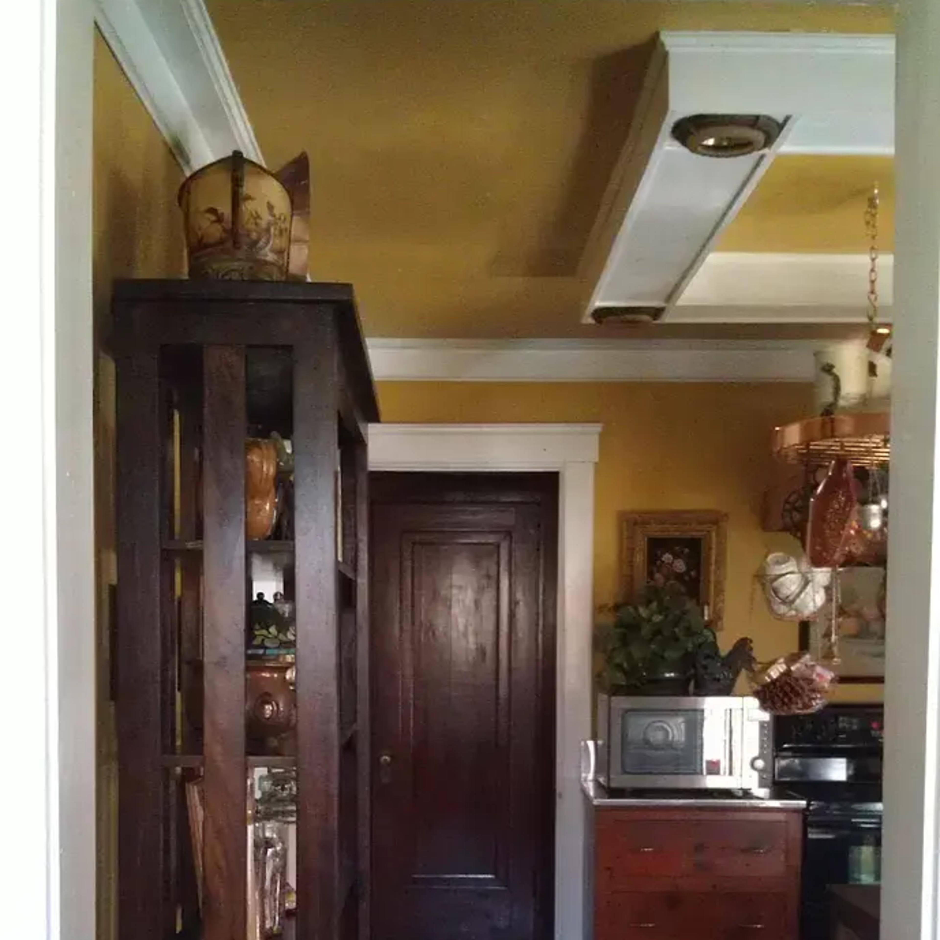 A narrow hallway leading to a door, with a wooden bookcase displaying various items on the left and a kitchenette area visible on the right.