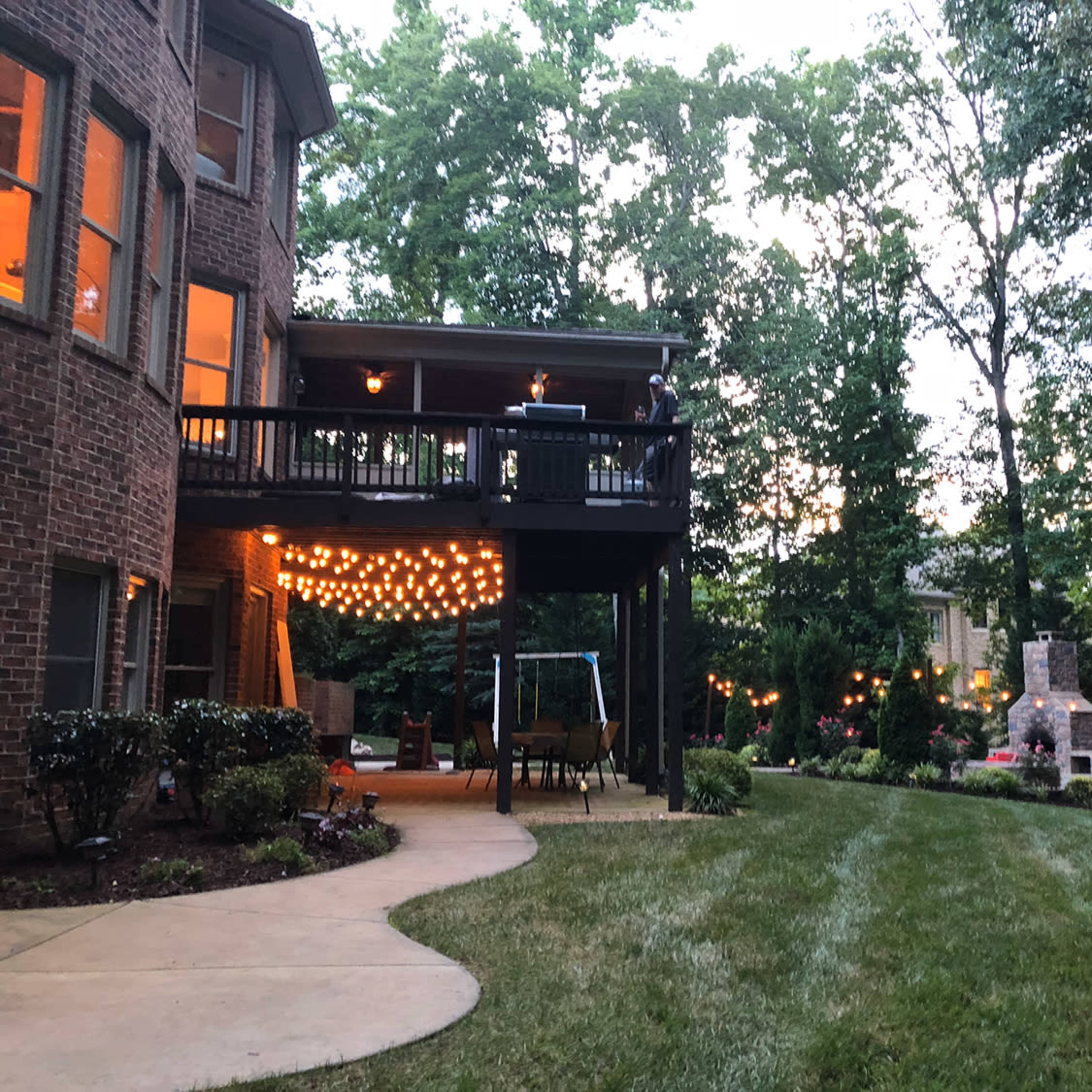 A well-lit backyard features a patio with a swing and a stone fireplace, surrounded by trees and lush grass.