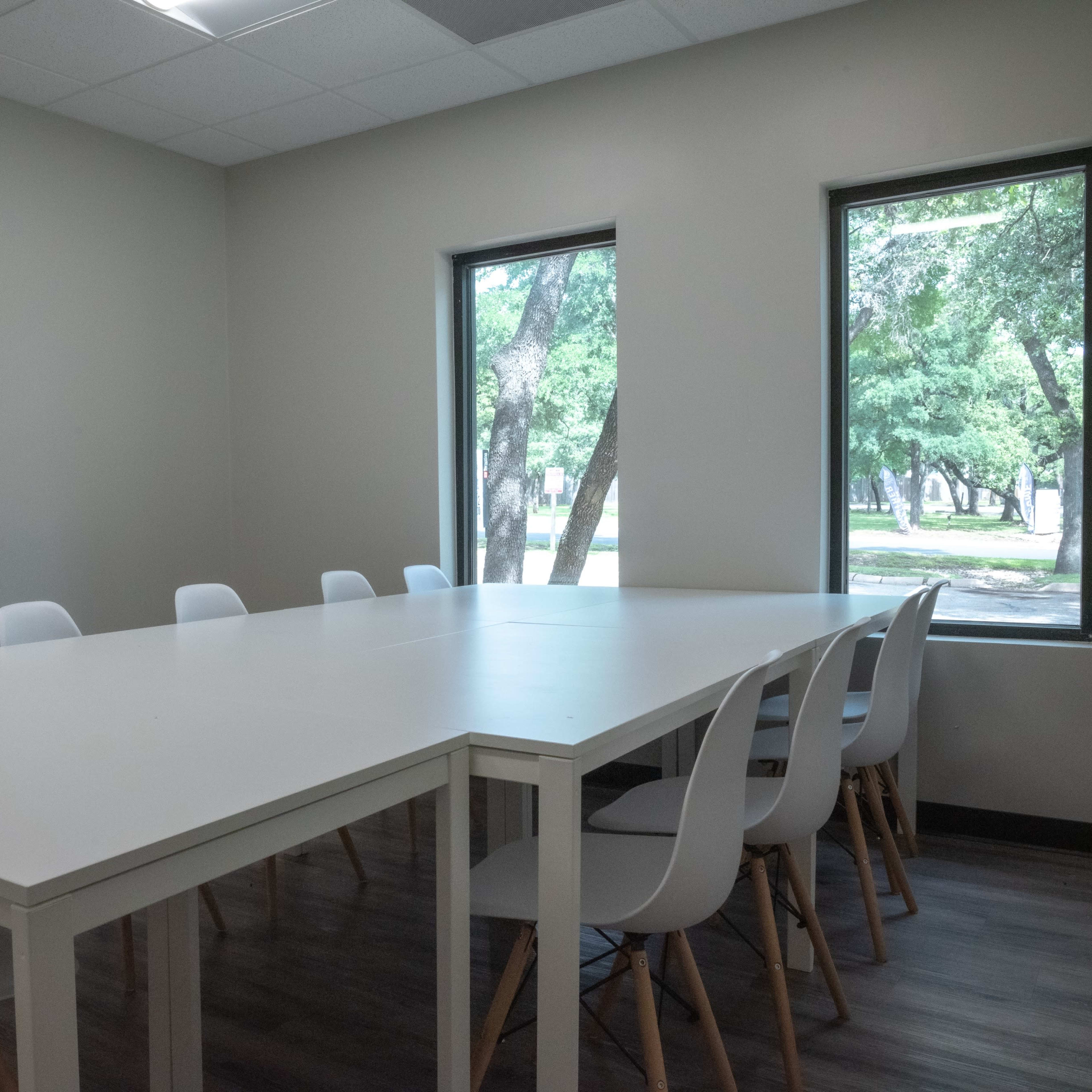 A spacious meeting room features a large white table surrounded by several chairs and is illuminated by natural light from two windows.