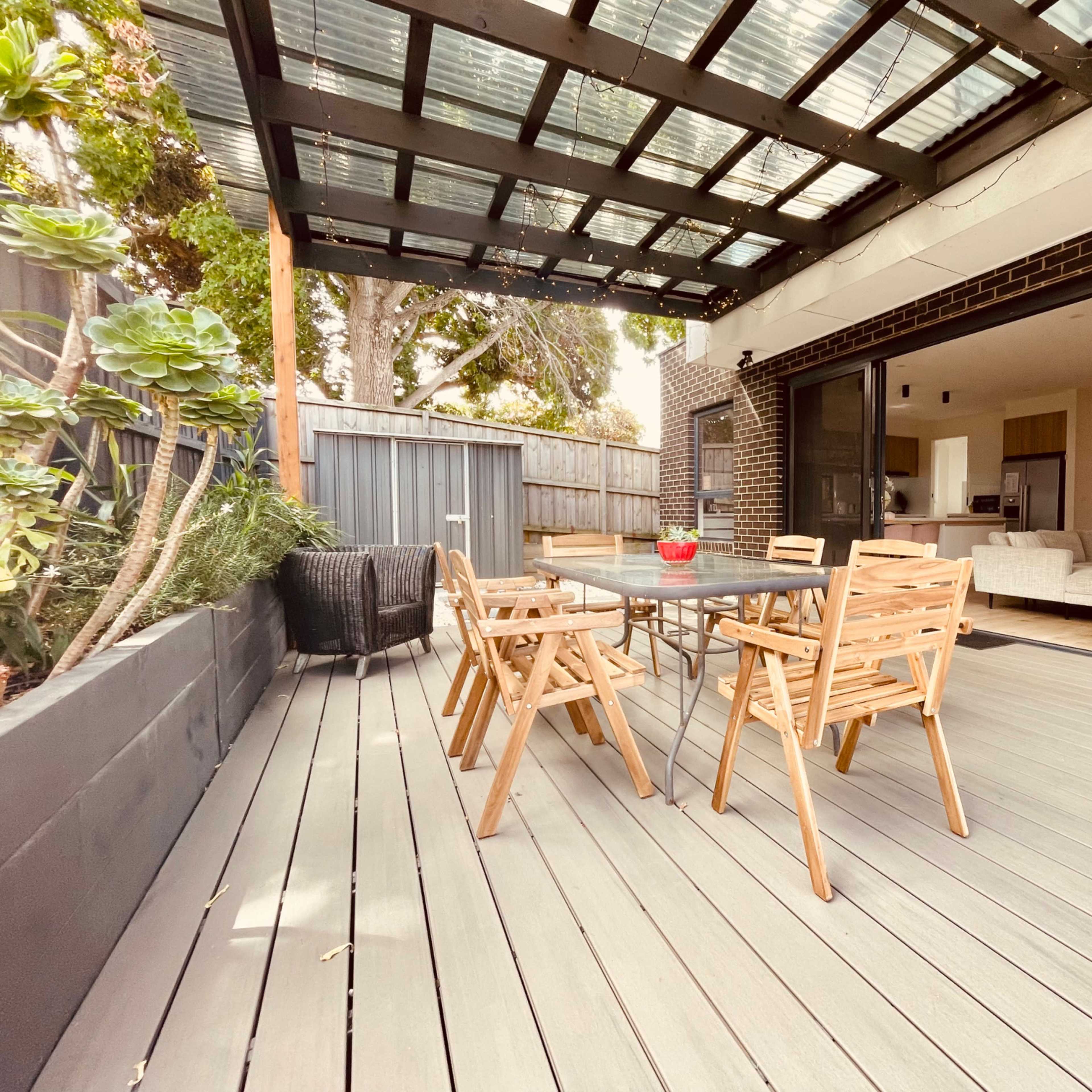 A wooden dining table surrounded by chairs is placed on a deck beneath a glass-covered patio.
