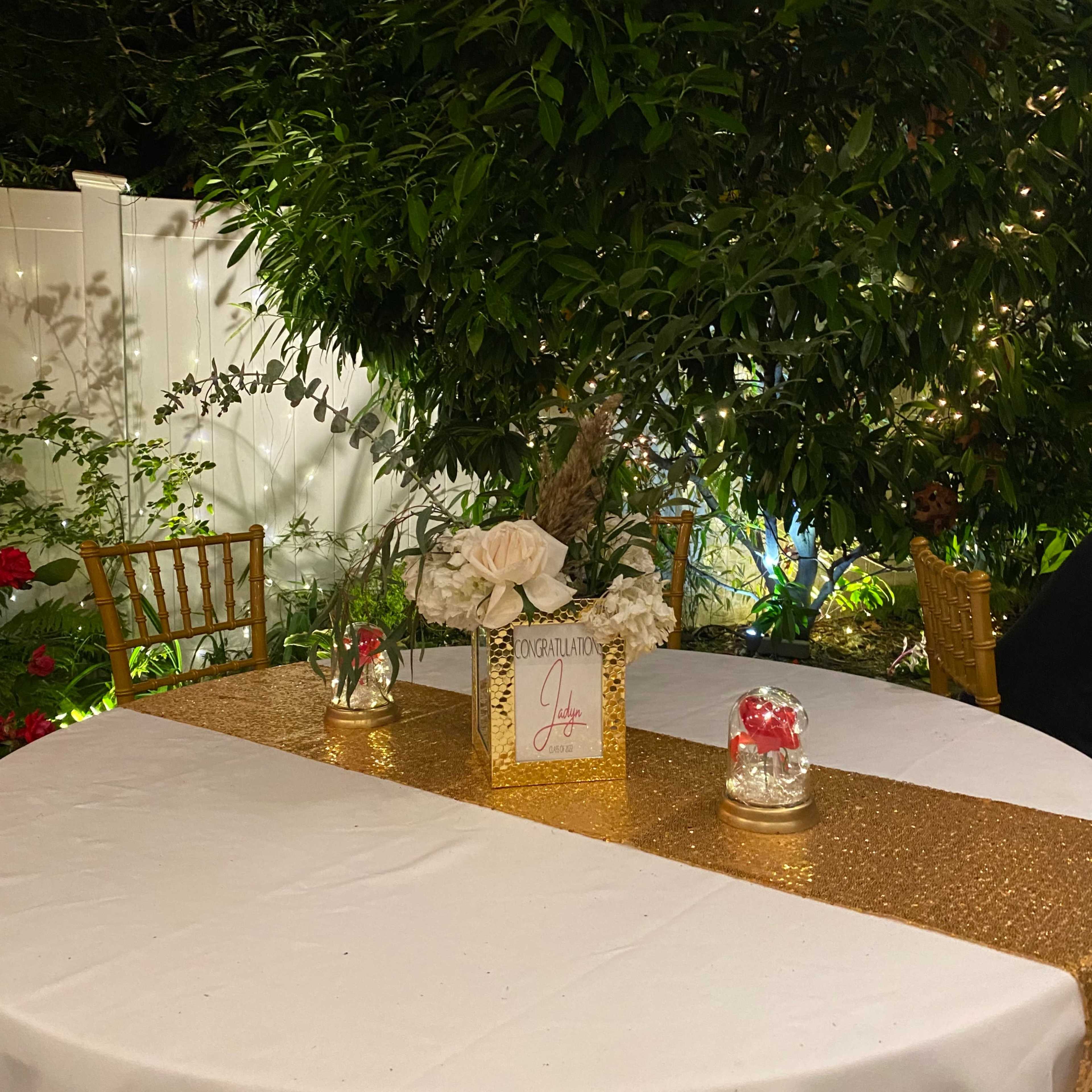 A neatly arranged table with a gold table runner, a floral centerpiece, and decorative candles is set in a garden surrounded by greenery and flowers.