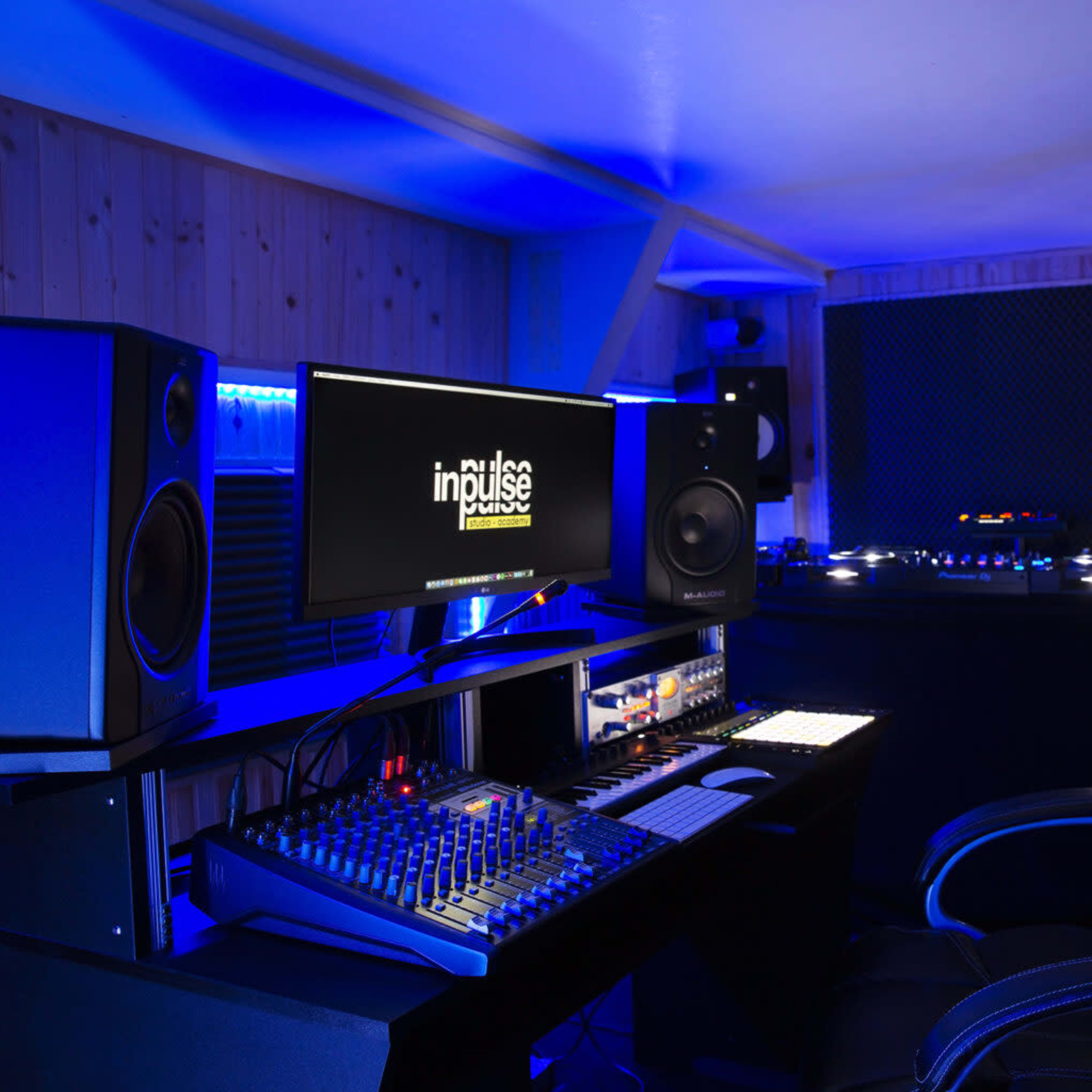 A recording studio features a central mixing console, large speakers, and a computer monitor displaying the logo "impulse," all illuminated by blue lighting.